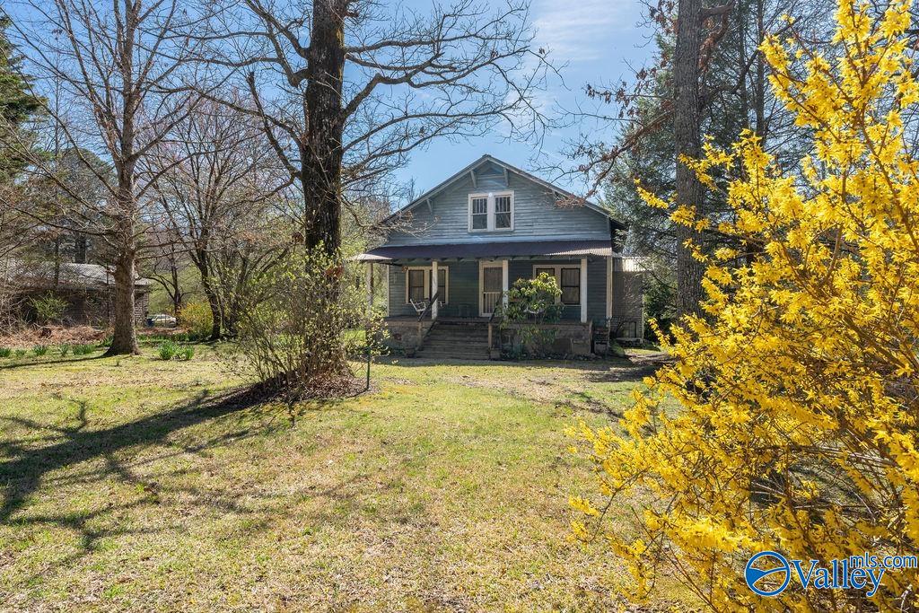 Gray-blue house with a covered porch in a grassy yard, framed by trees and a bright yellow shrub.