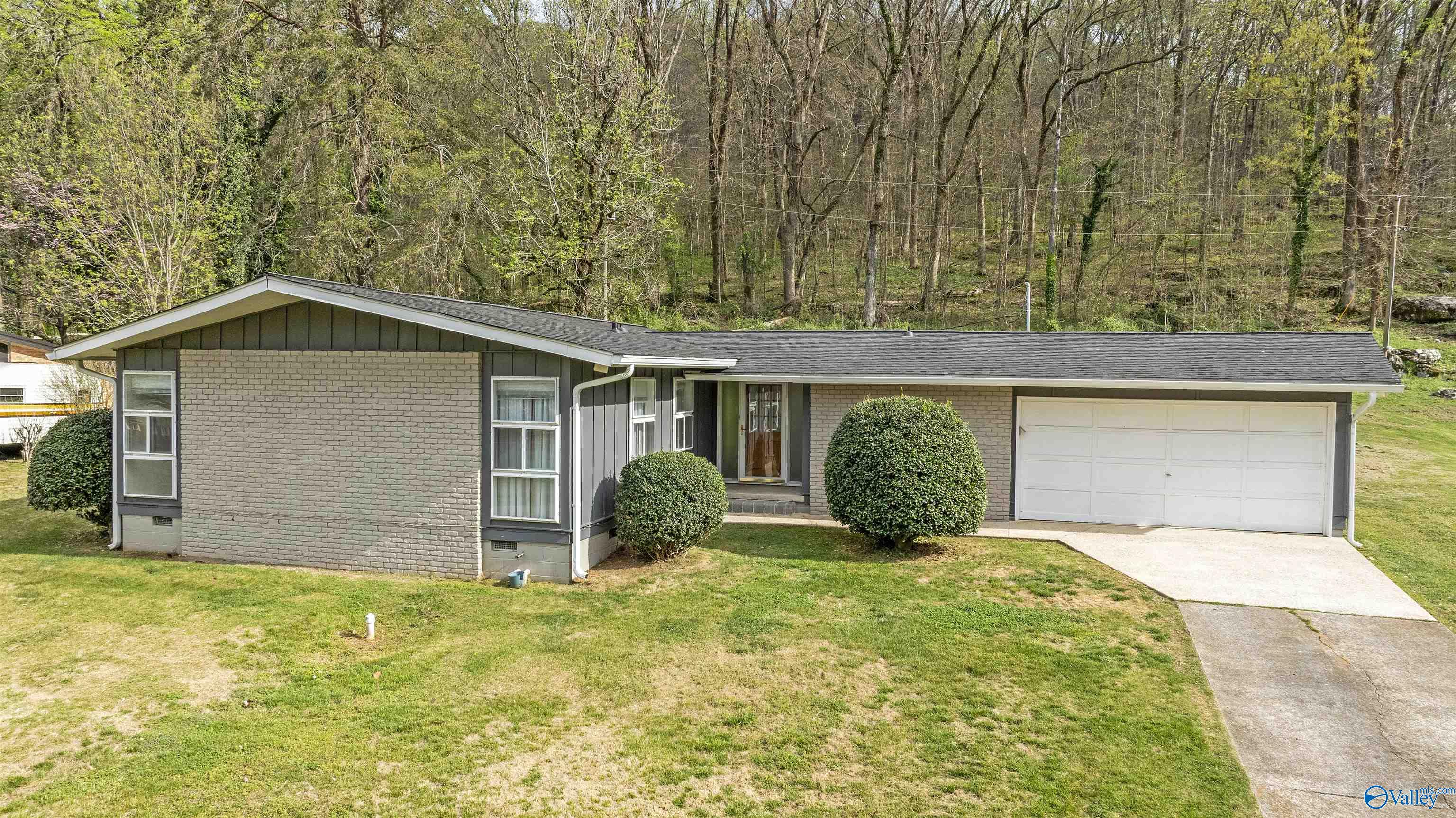 Single-story brick house with an attached two-car garage and wooded hillside behind it.