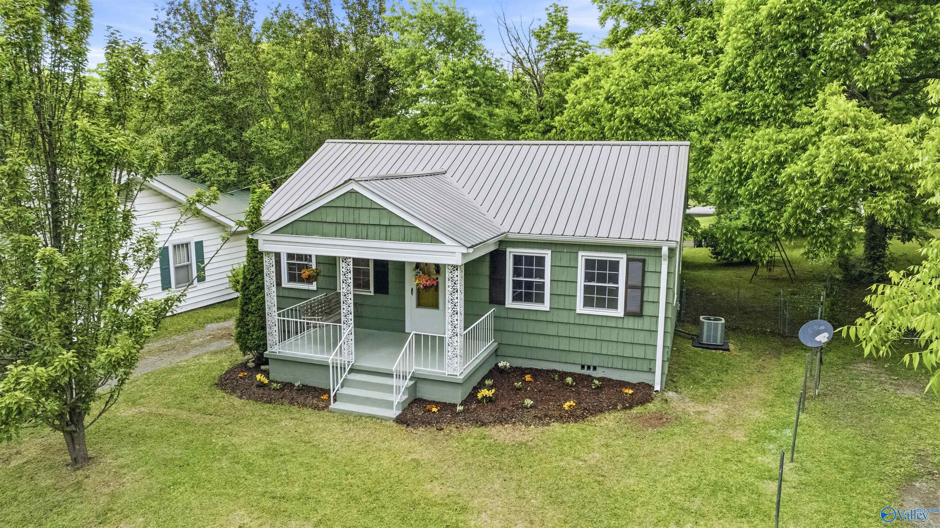Green house with a light metal roof, white porch, and landscaped yard among mature trees.