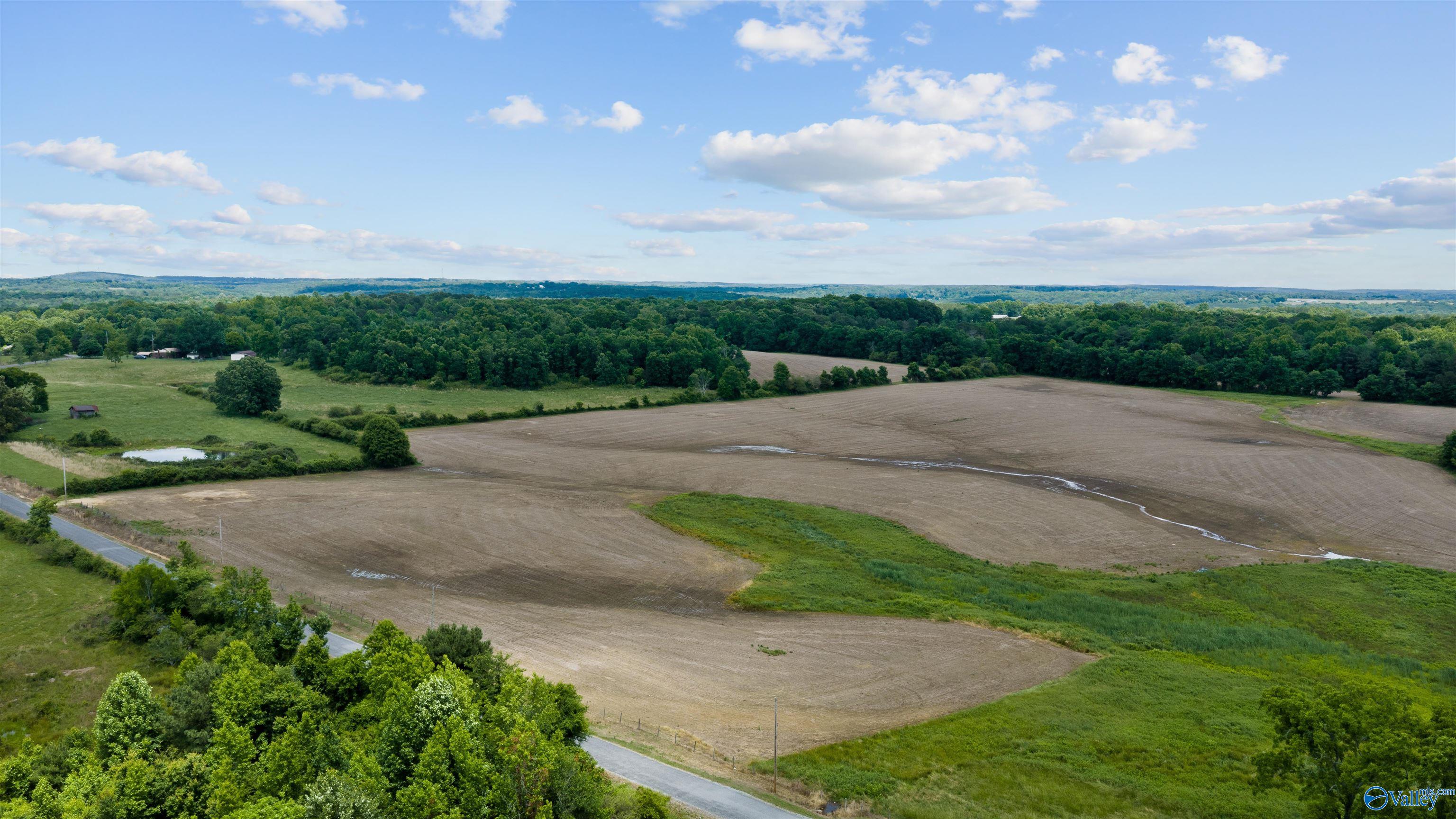 Aerial view of rural acreage with plowed fields, green pasture, a small pond, and a country road.