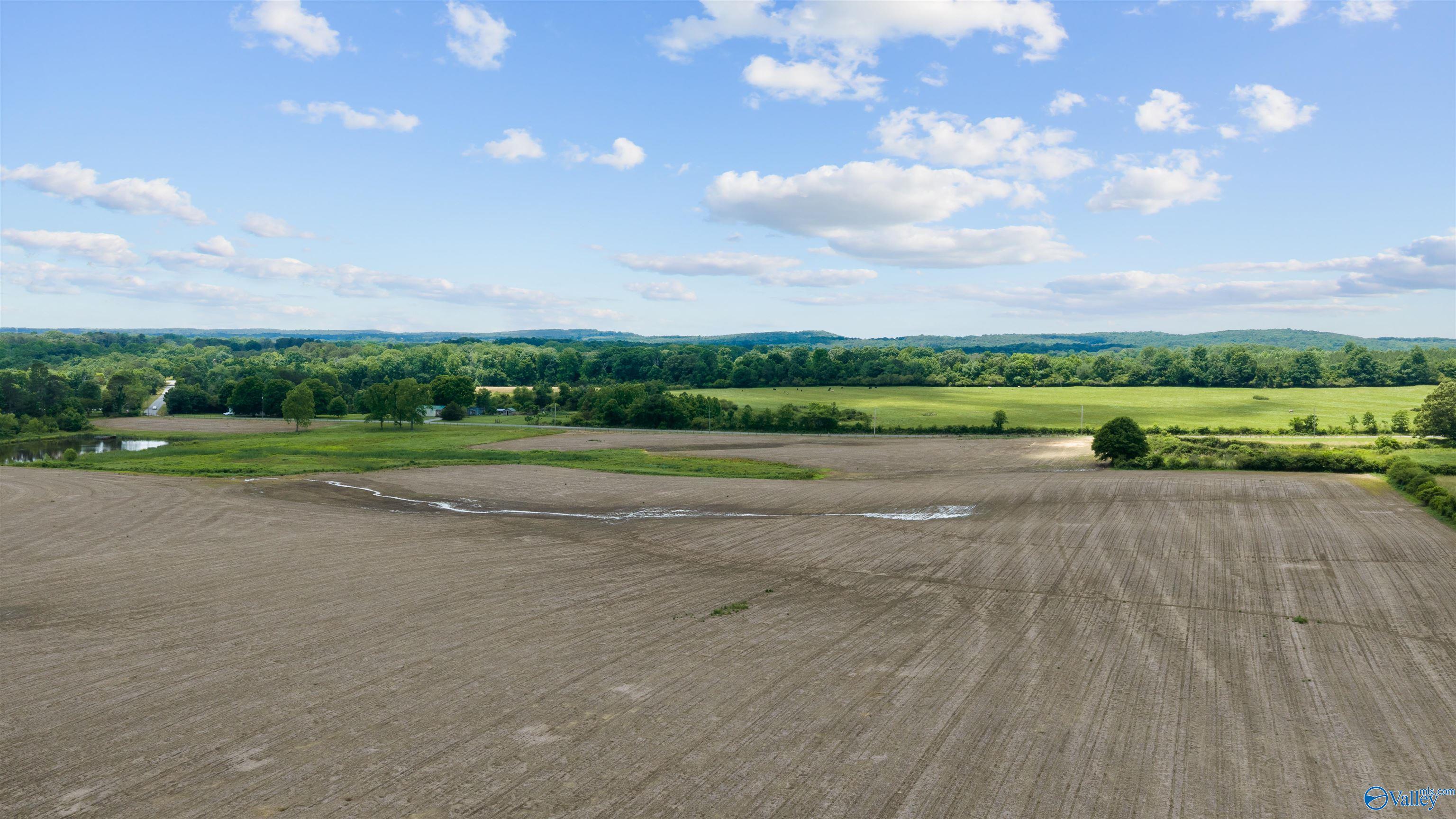 Aerial view of tilled acreage, a small pond, green pasture, and wooded hills.