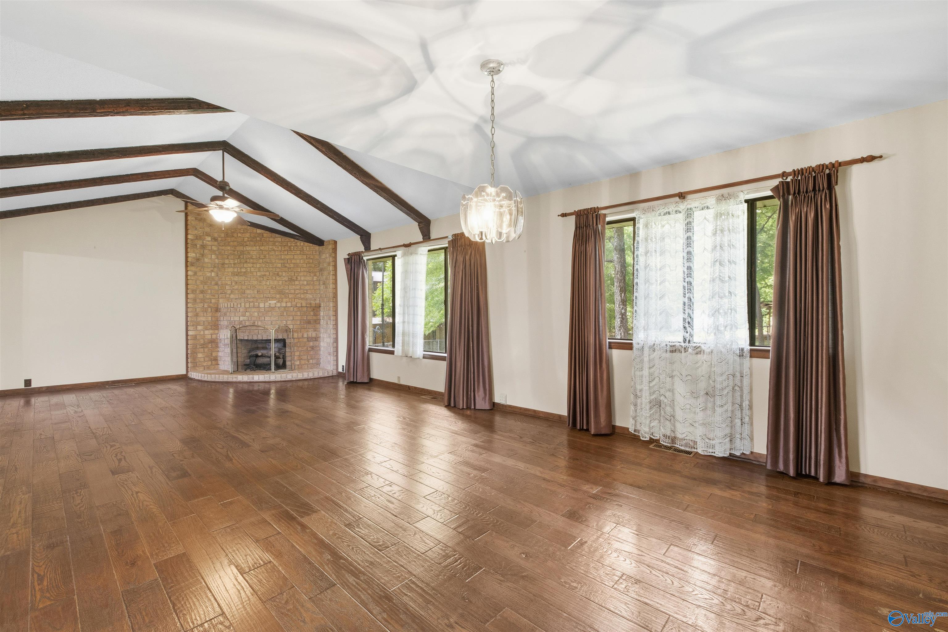 Vacant living room with vaulted beam ceiling, brick fireplace, and brown wood-look floors.