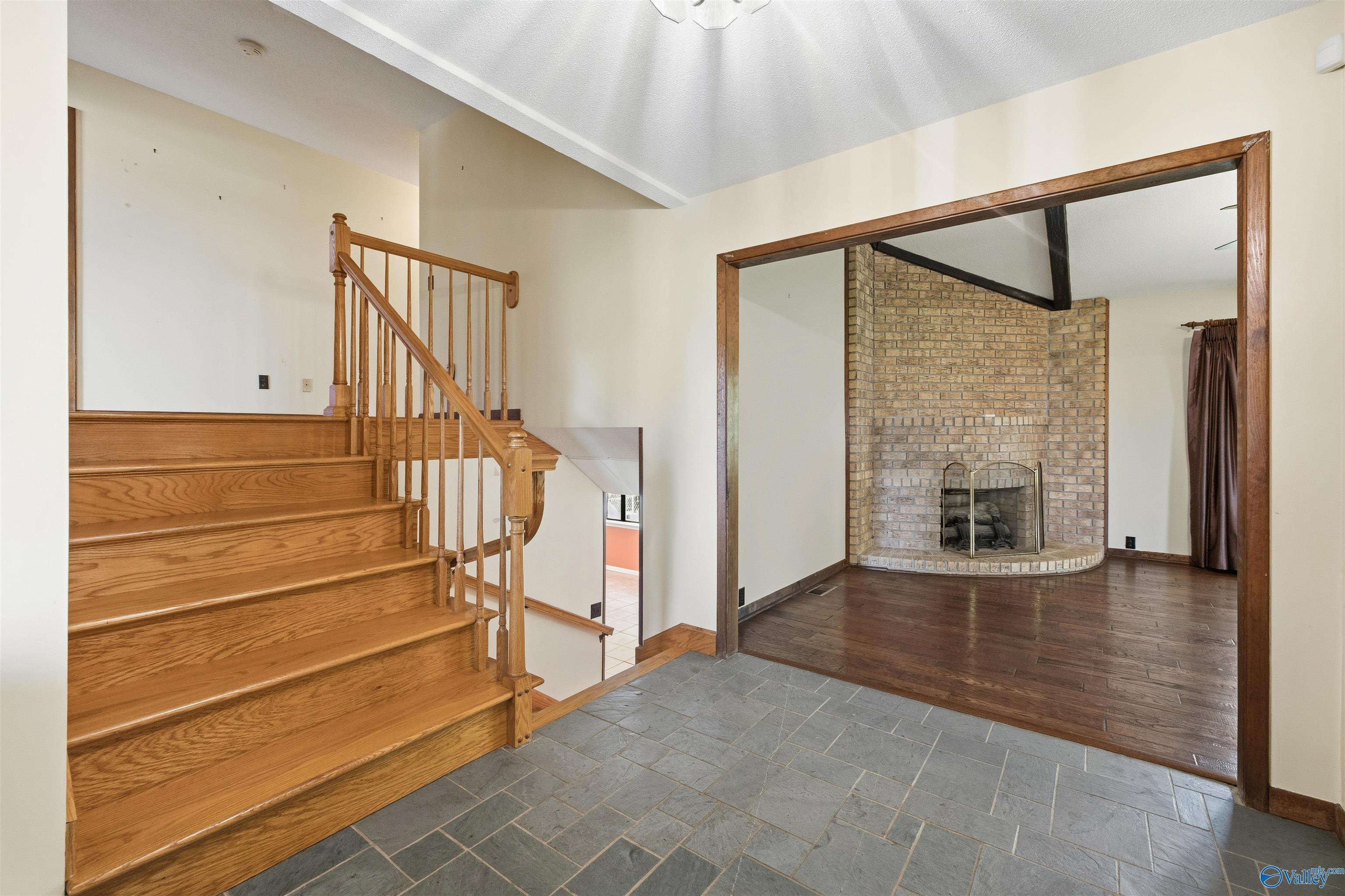 Home entryway with a gray tile floor and wooden staircase beside a living room with a brick fireplace.