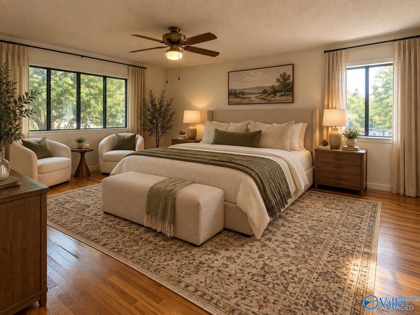 Staged bedroom with a large upholstered bed, two bright windows, armchairs, and a patterned rug.