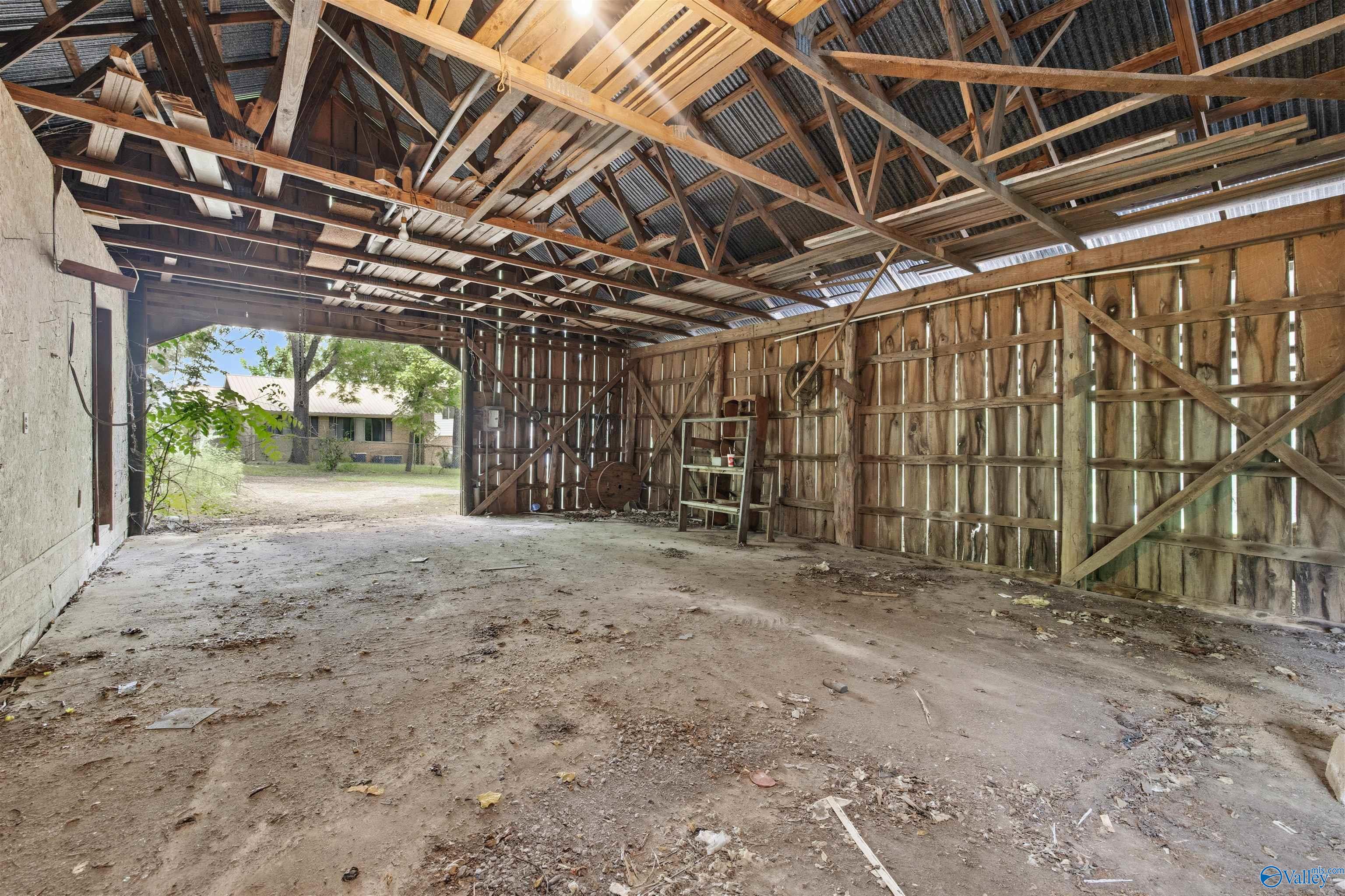 Empty barn interior with exposed timber rafters, corrugated roof, and an open bay facing a house.