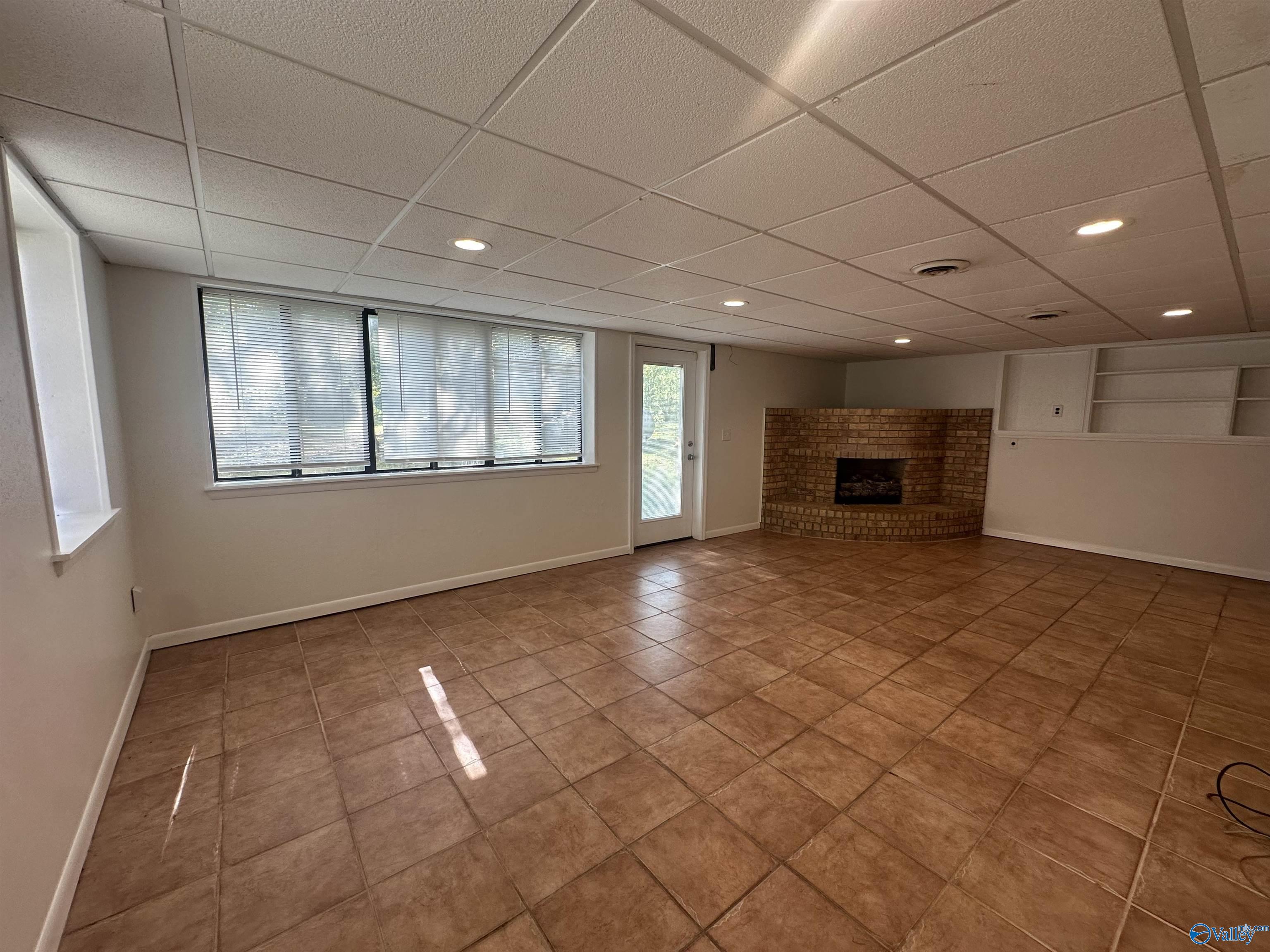 Vacant room with brown tile flooring, wide windows, built-in shelving, and a brick fireplace.