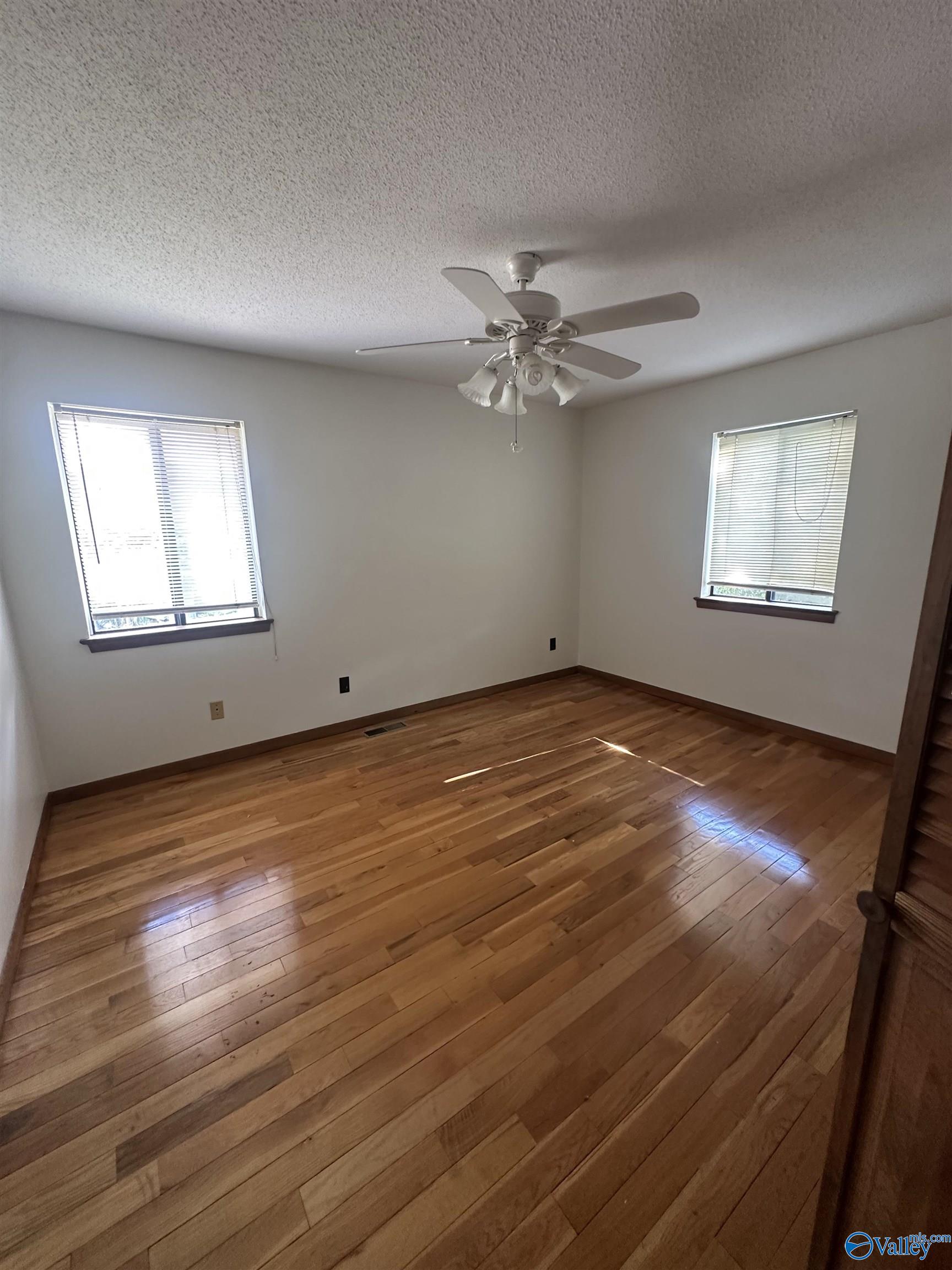 Empty bedroom with polished hardwood floors, two windows with blinds, and a ceiling fan.