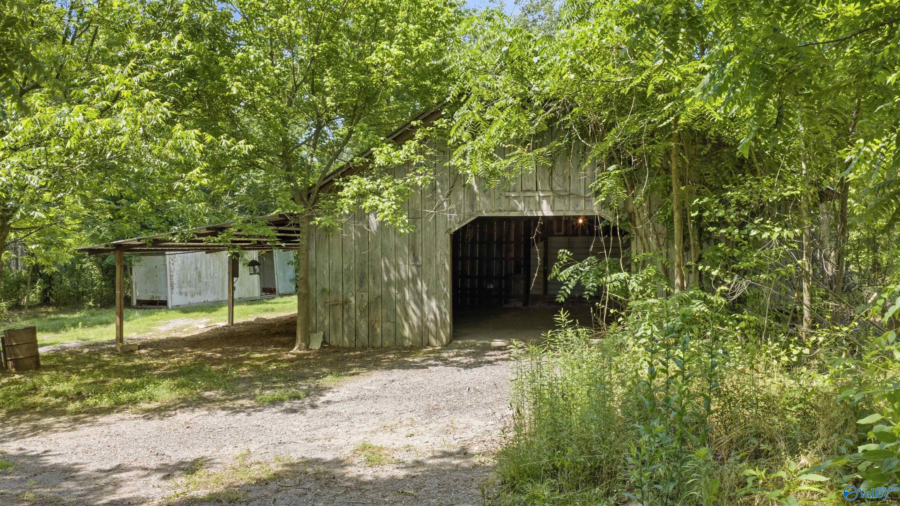 Weathered wooden barn with an open bay and lean-to beneath dense trees beside a gravel drive.