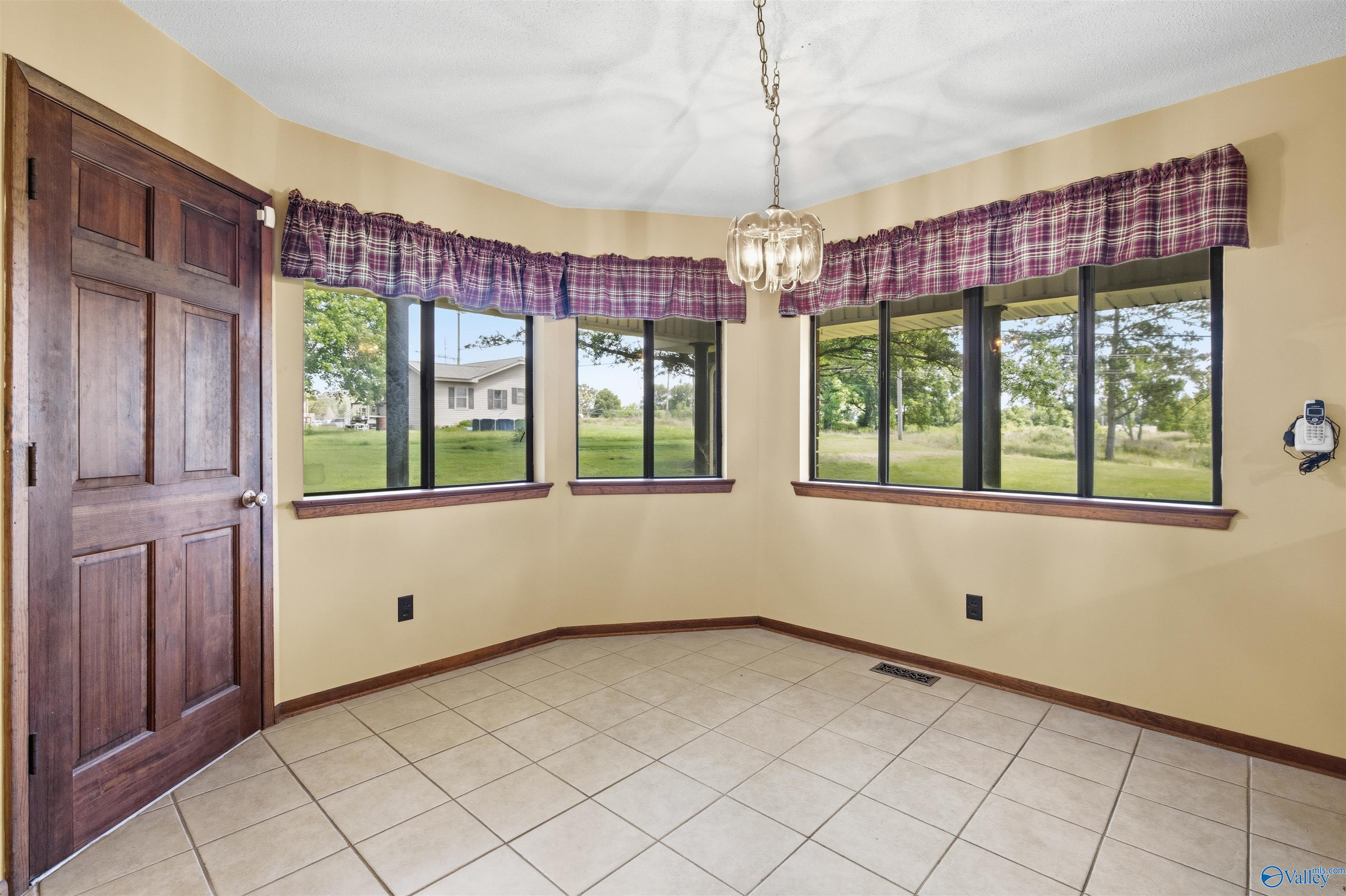 Empty tiled room with a dark wood door, broad windows, and purple plaid valances.