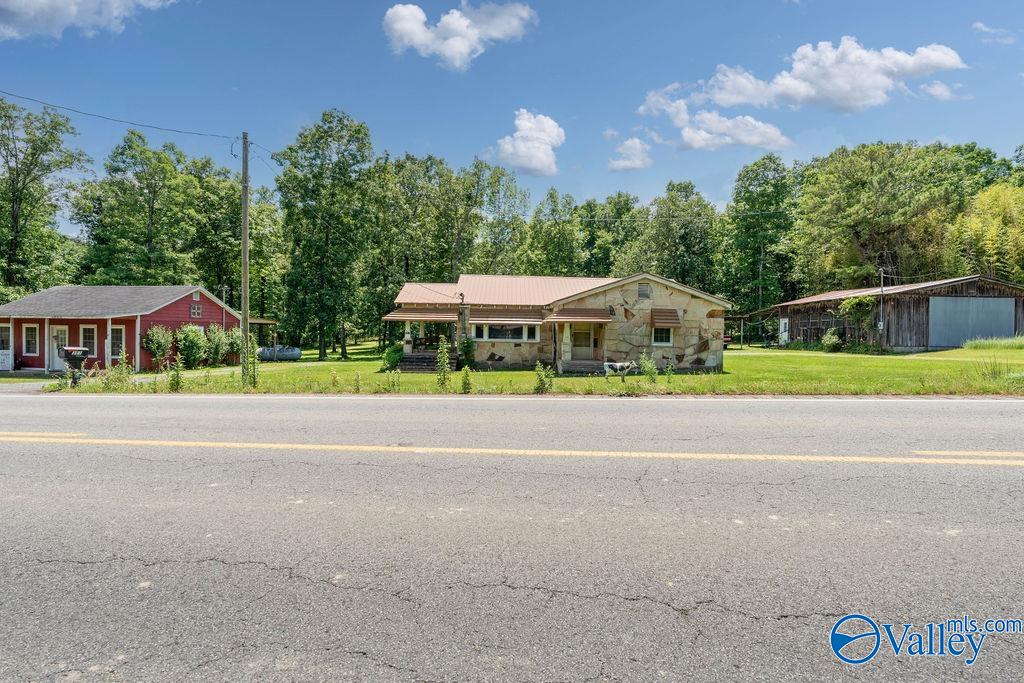 Stone-front home with a covered porch, red outbuilding, and barn across a two-lane road.