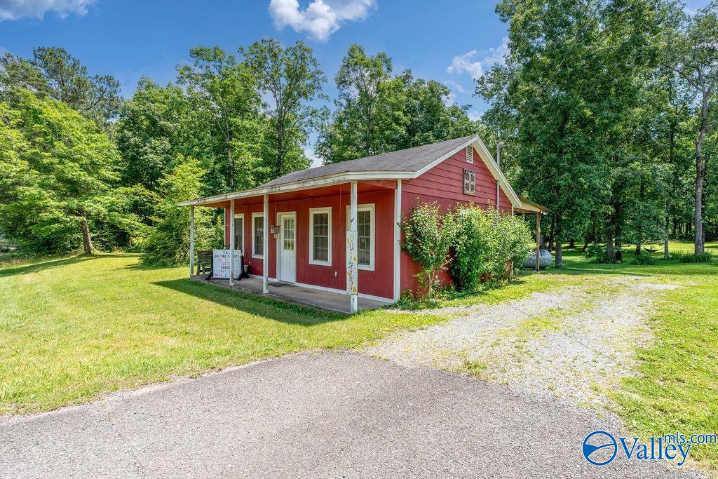 Small red building with a covered porch on a grassy, wooded lot beside a gravel drive.