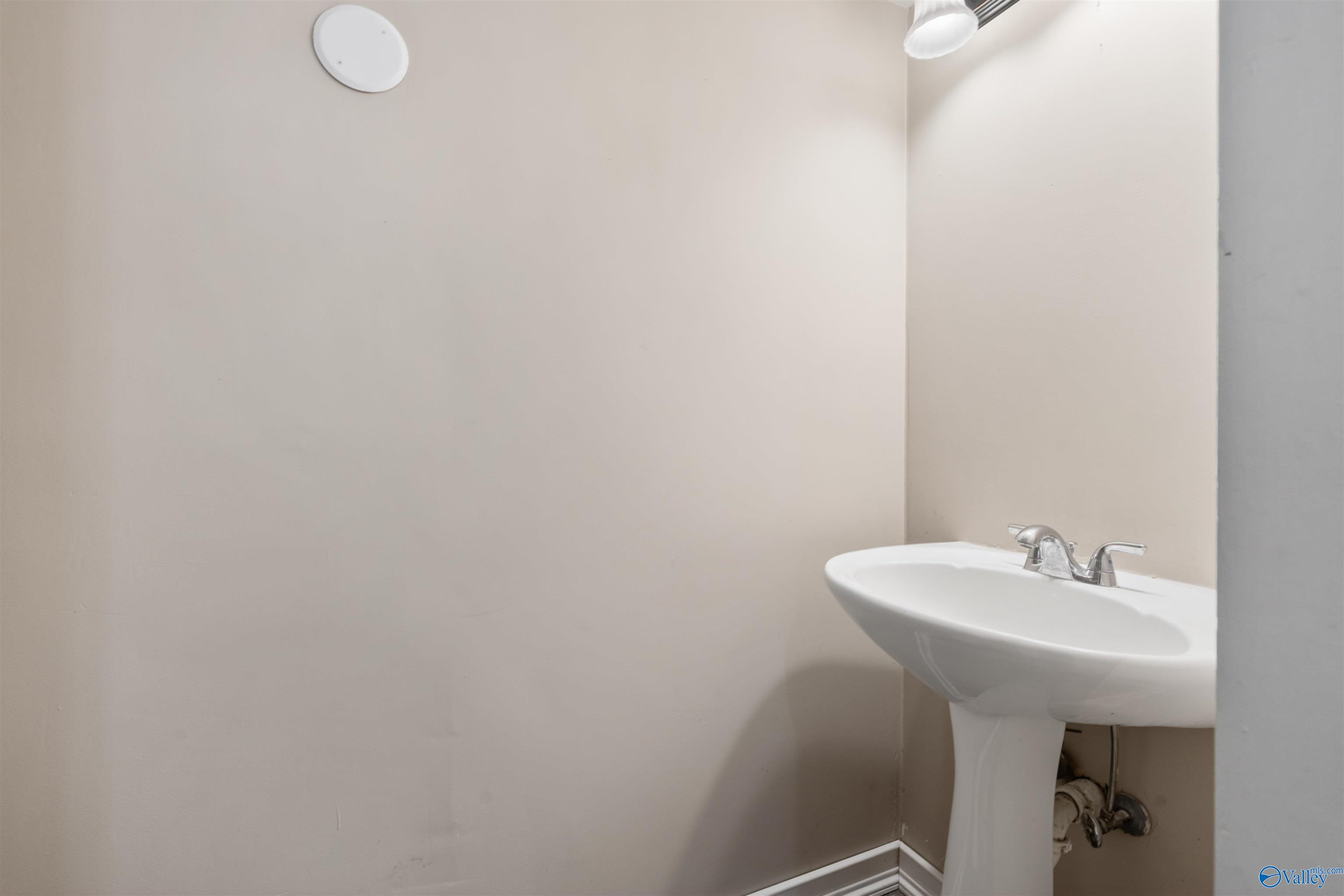 Bathroom pedestal sink with chrome faucets against beige walls and exposed plumbing.