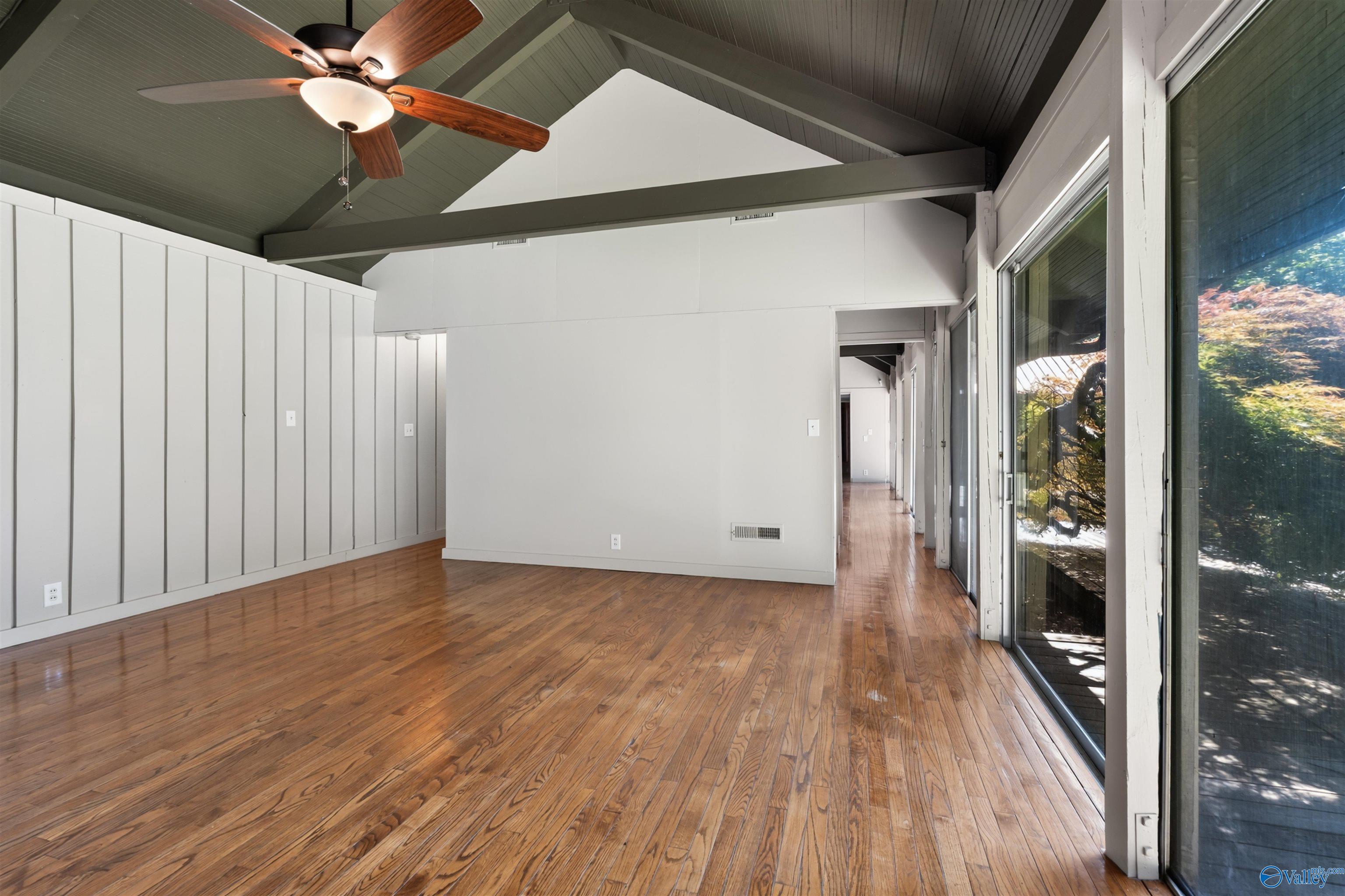 Empty room with polished hardwood floors, vaulted beamed ceiling, ceiling fan, and tall windows overlooking trees.
