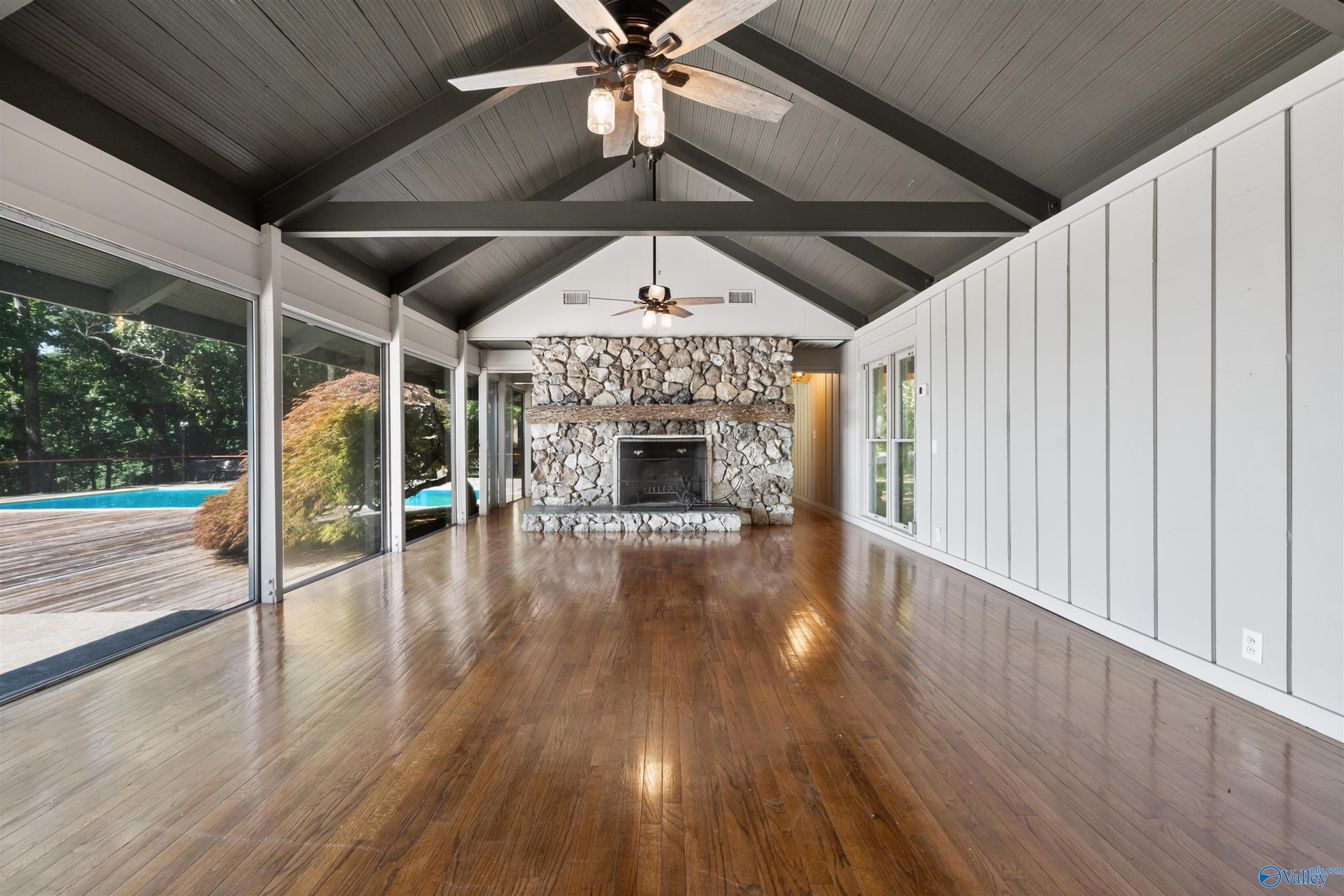 Living room with a vaulted beamed ceiling, stone fireplace, hardwood floors, and sliding doors overlooking a pool.