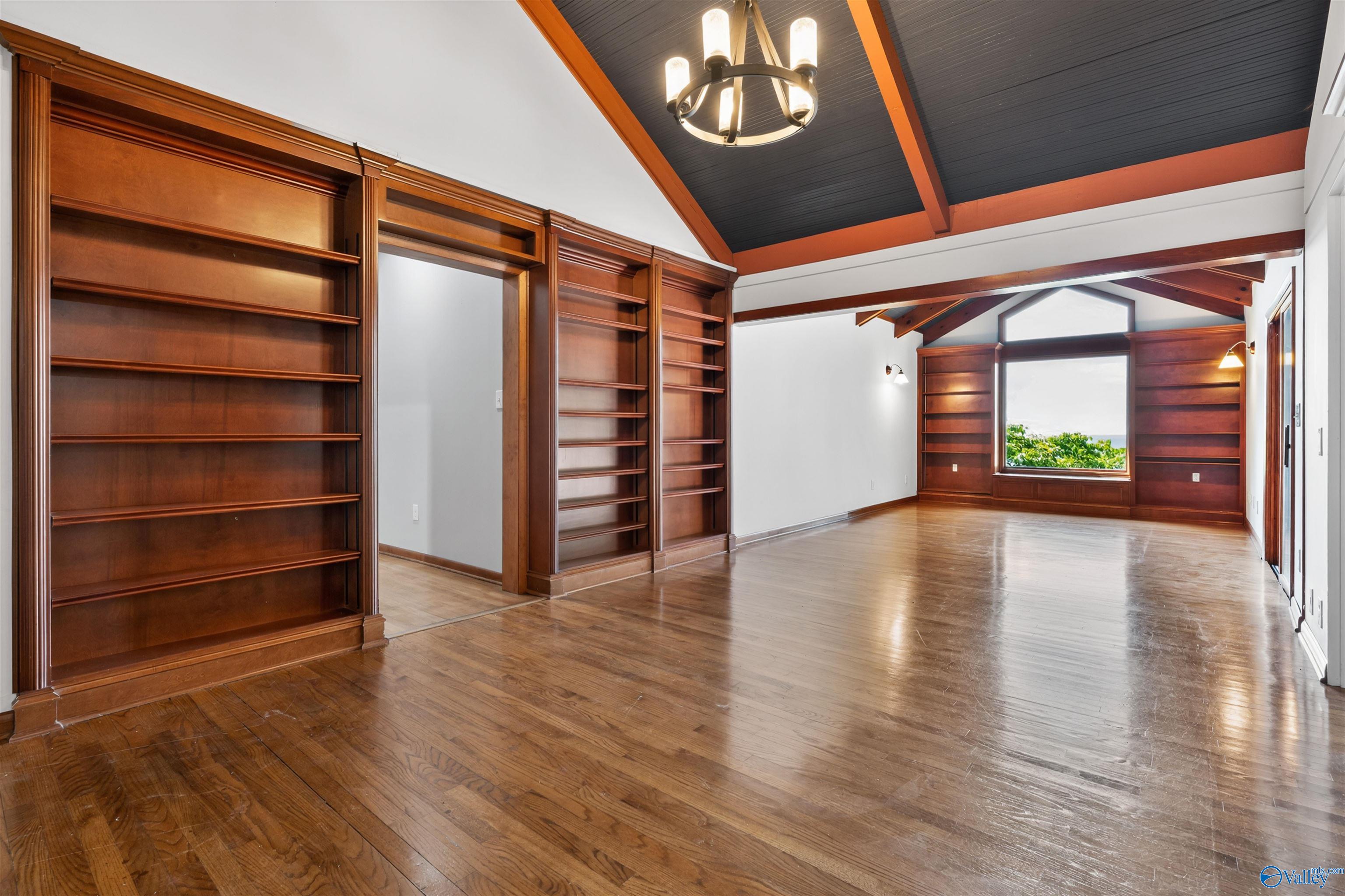 Empty room with built-in wooden bookshelves, polished floors, vaulted ceiling, and a large window.