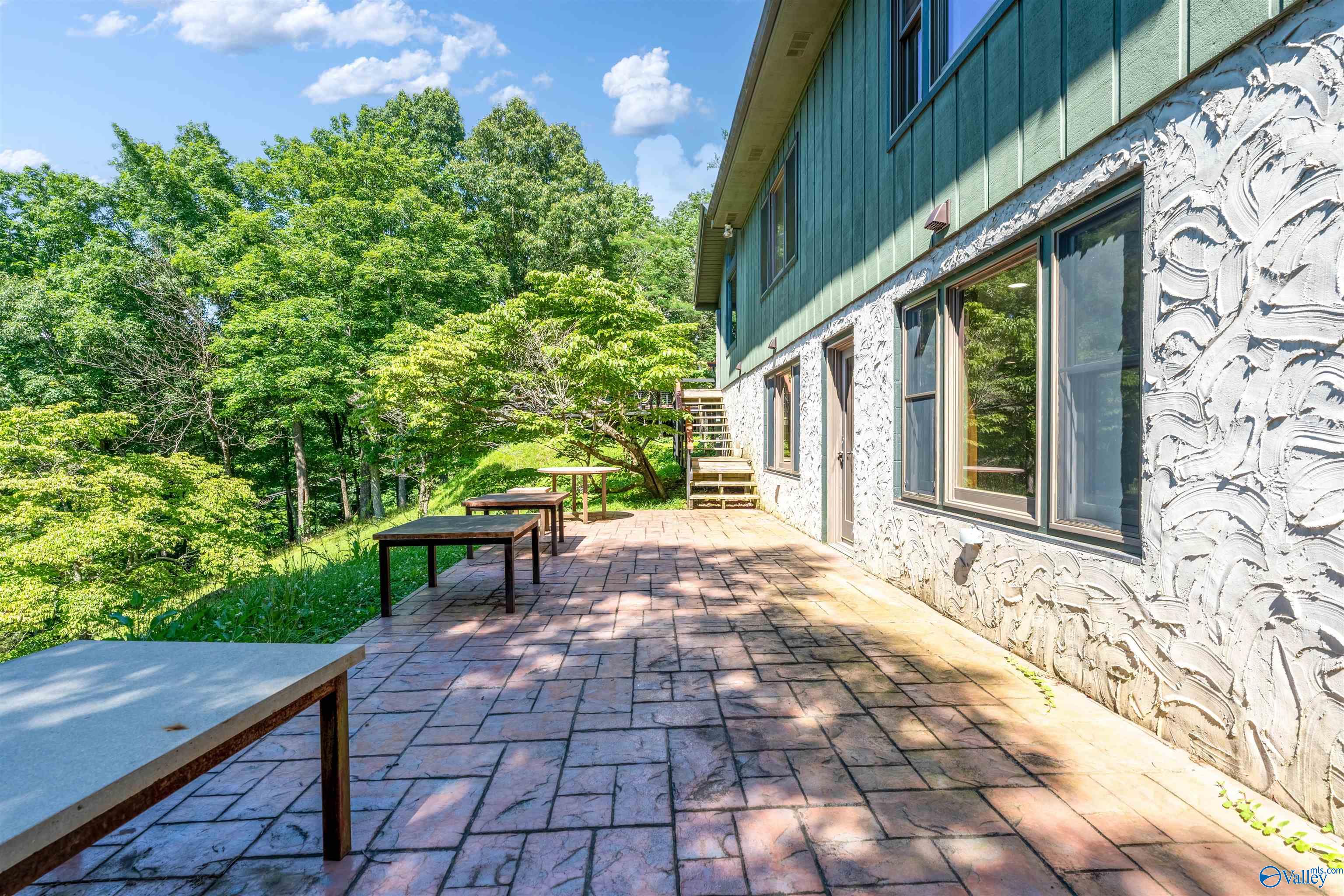 Stone-paved patio beside a green-sided home, with outdoor tables and a wooded slope.