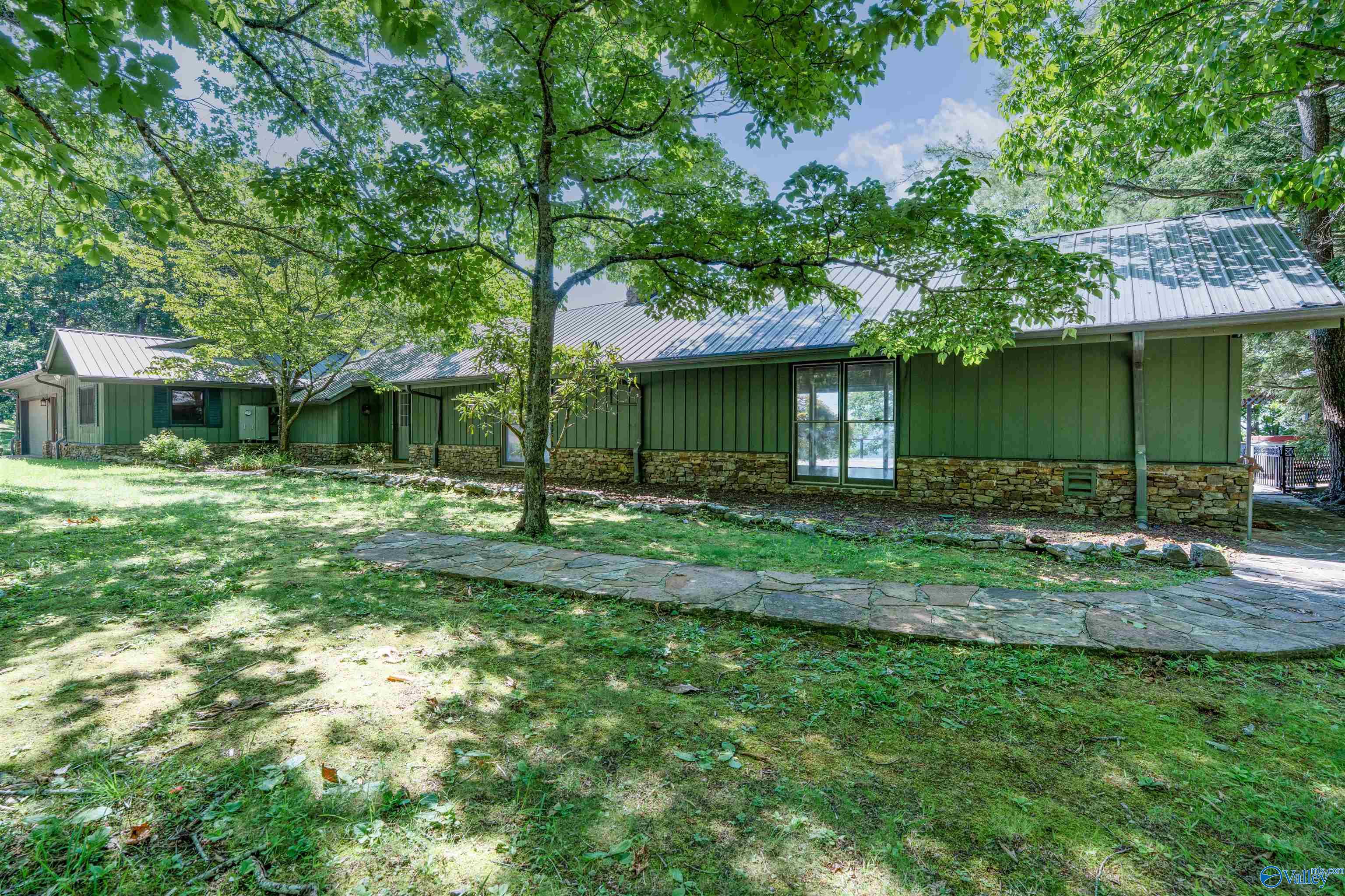 Green single-story home with stone base and metal roof, shaded by mature trees beside a flagstone walkway.