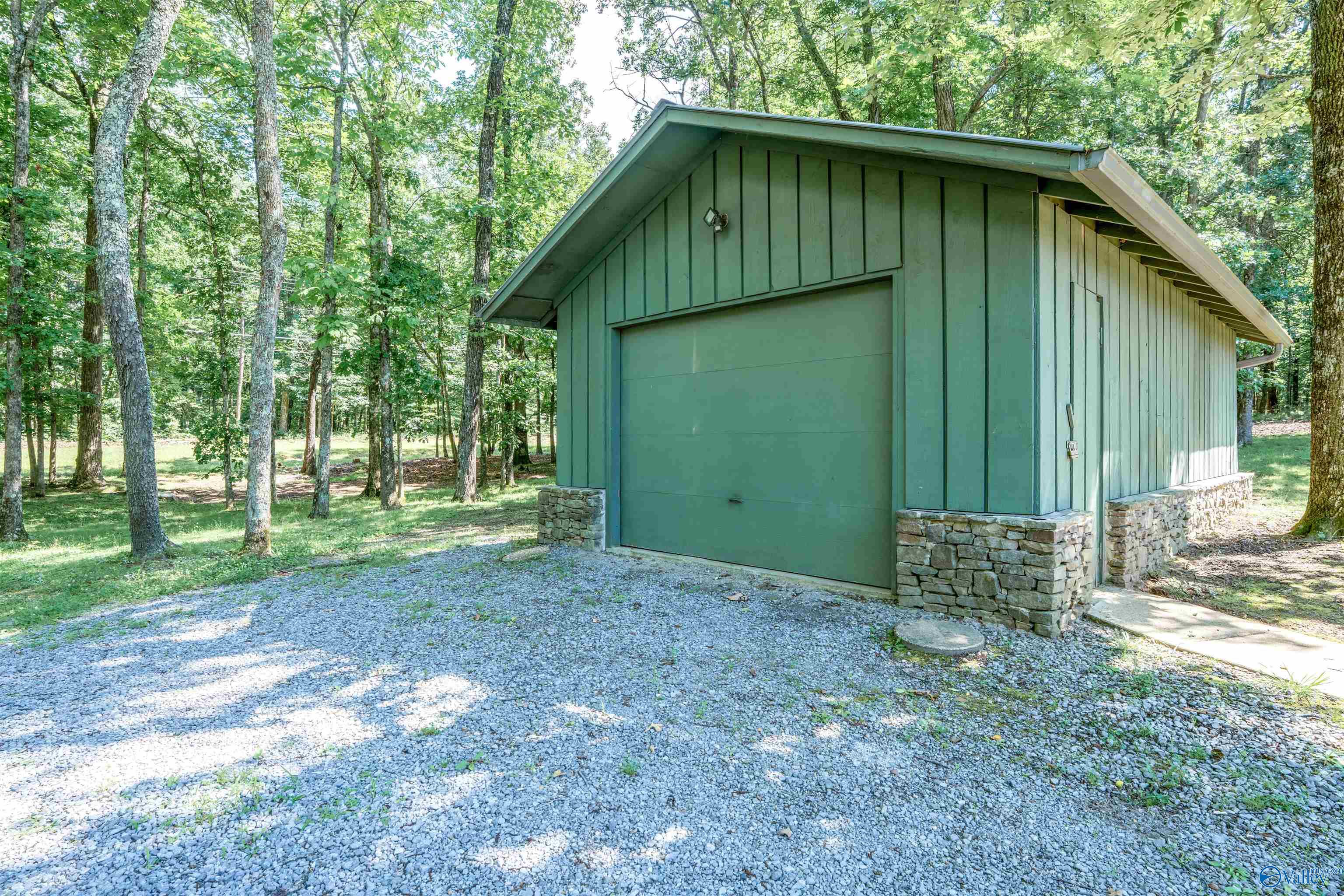 Green detached garage with a stone base, surrounded by trees beside a gravel driveway.