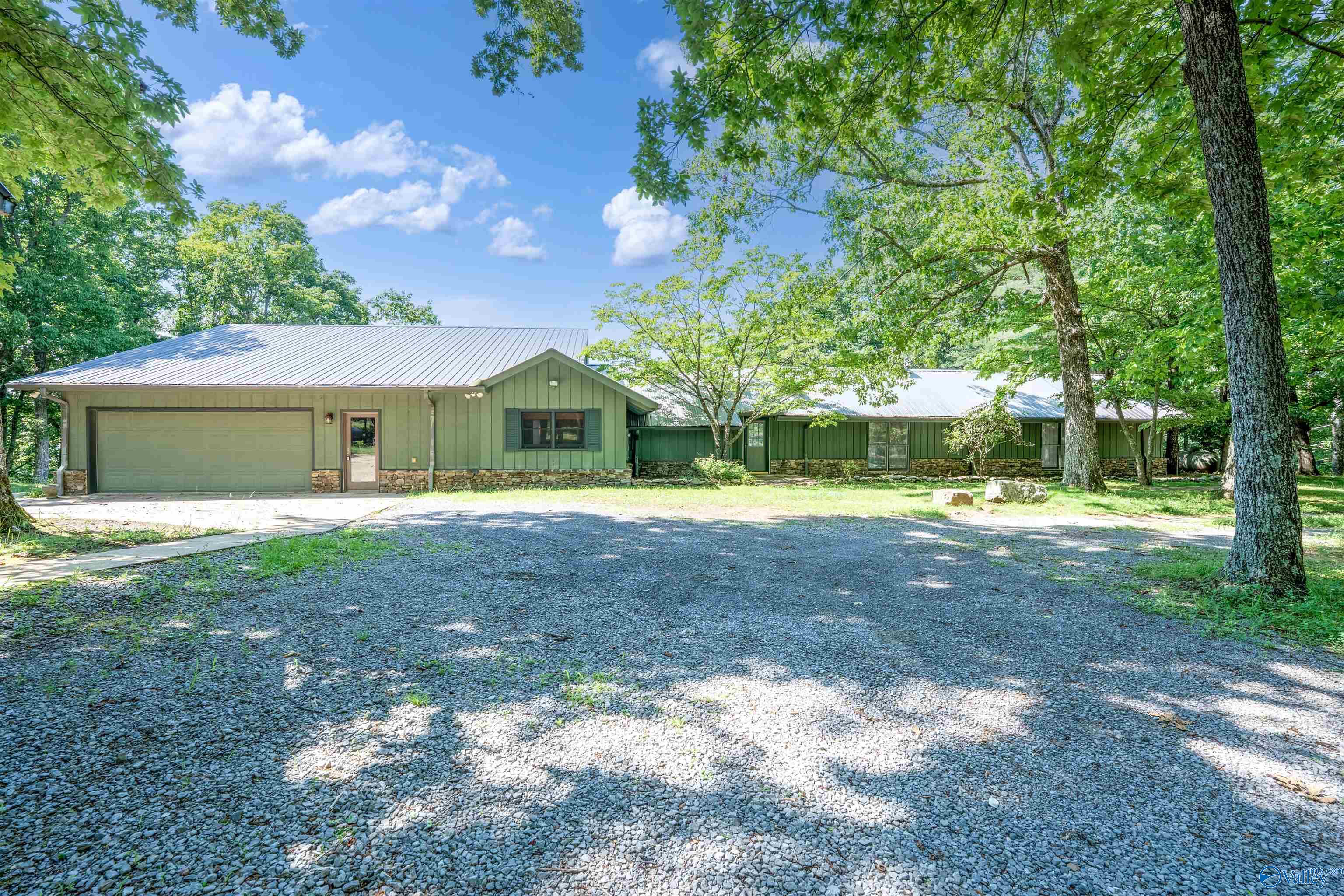 Green single-story home with an attached garage, stone accents, and a gravel driveway beneath mature trees.