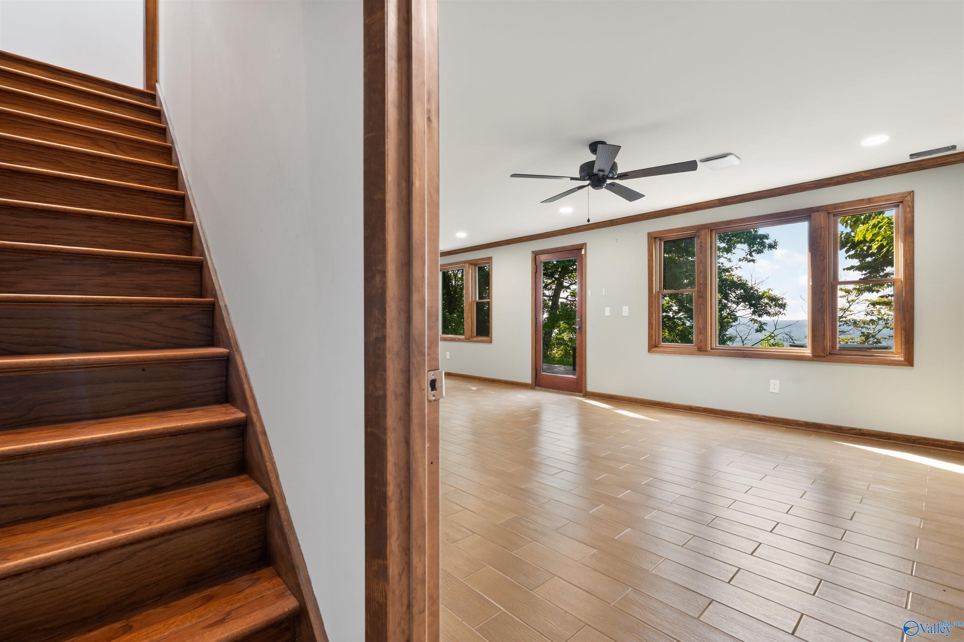 Empty room with wood-trimmed windows, tiled floor, ceiling fan, and dark wood staircase.