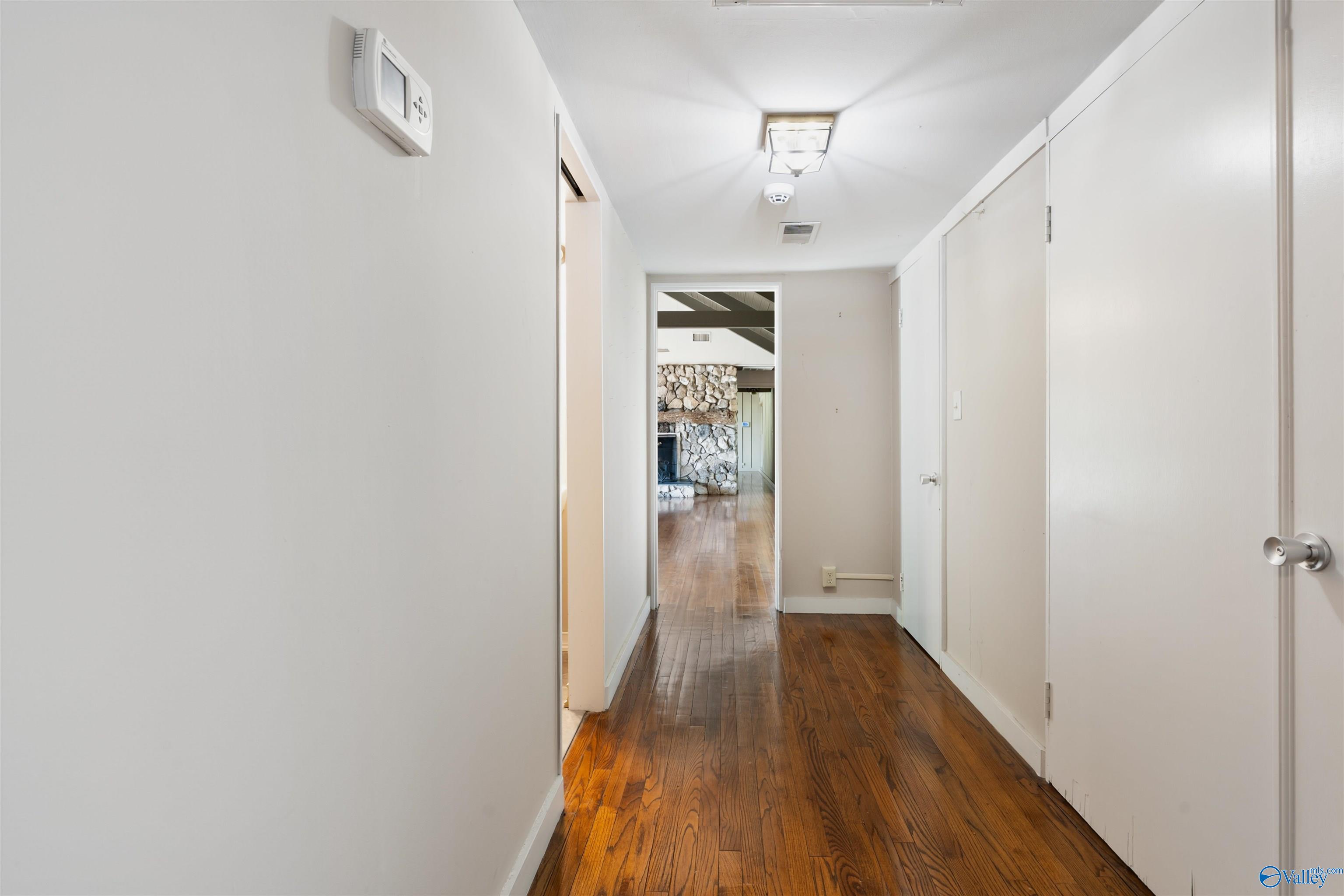Empty hallway with polished hardwood floors leading to a room with a stone fireplace.