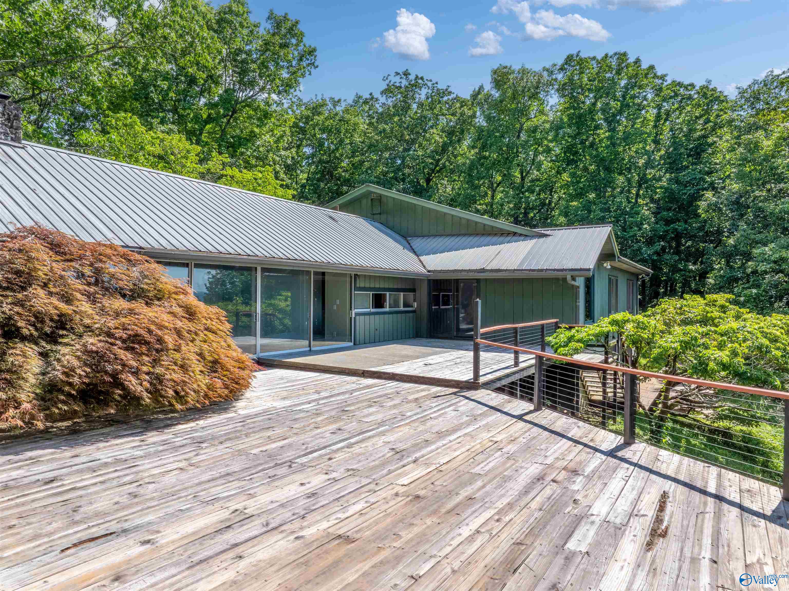 Green home with a metal roof and expansive wooden deck, surrounded by trees.