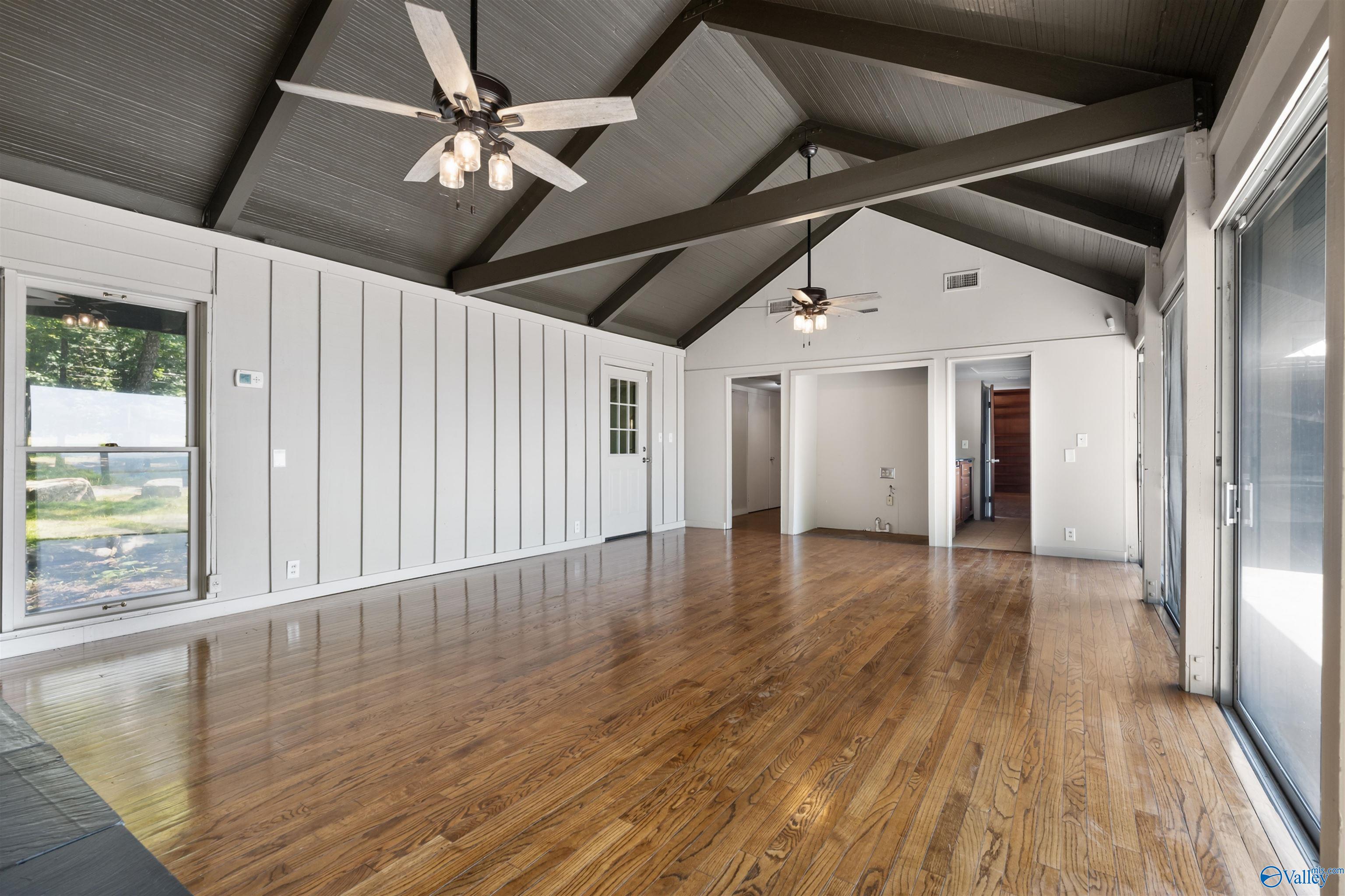 Spacious vacant living room with hardwood floors, vaulted beam ceiling, ceiling fans, and large sliding glass doors.