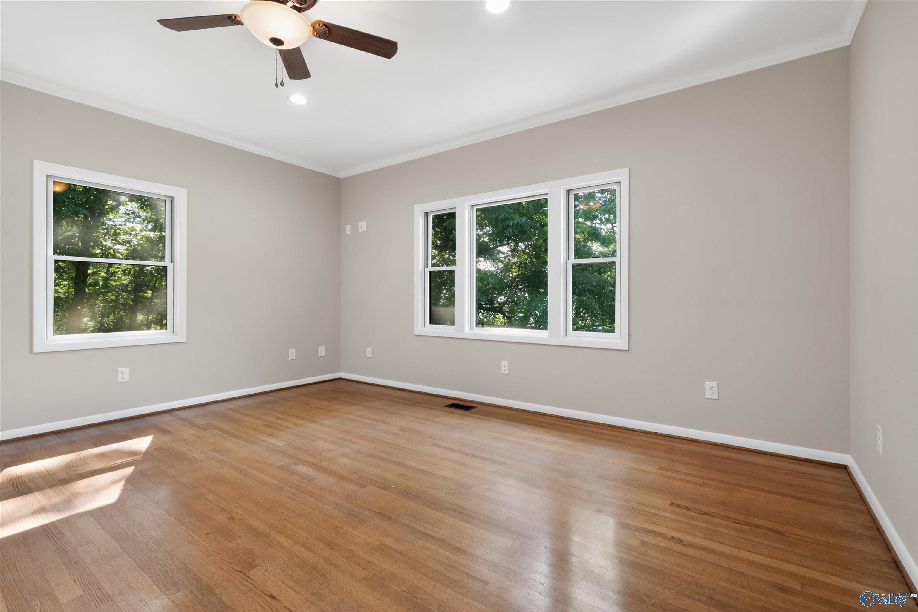 Vacant room with hardwood floors, a ceiling fan, and windows overlooking trees.