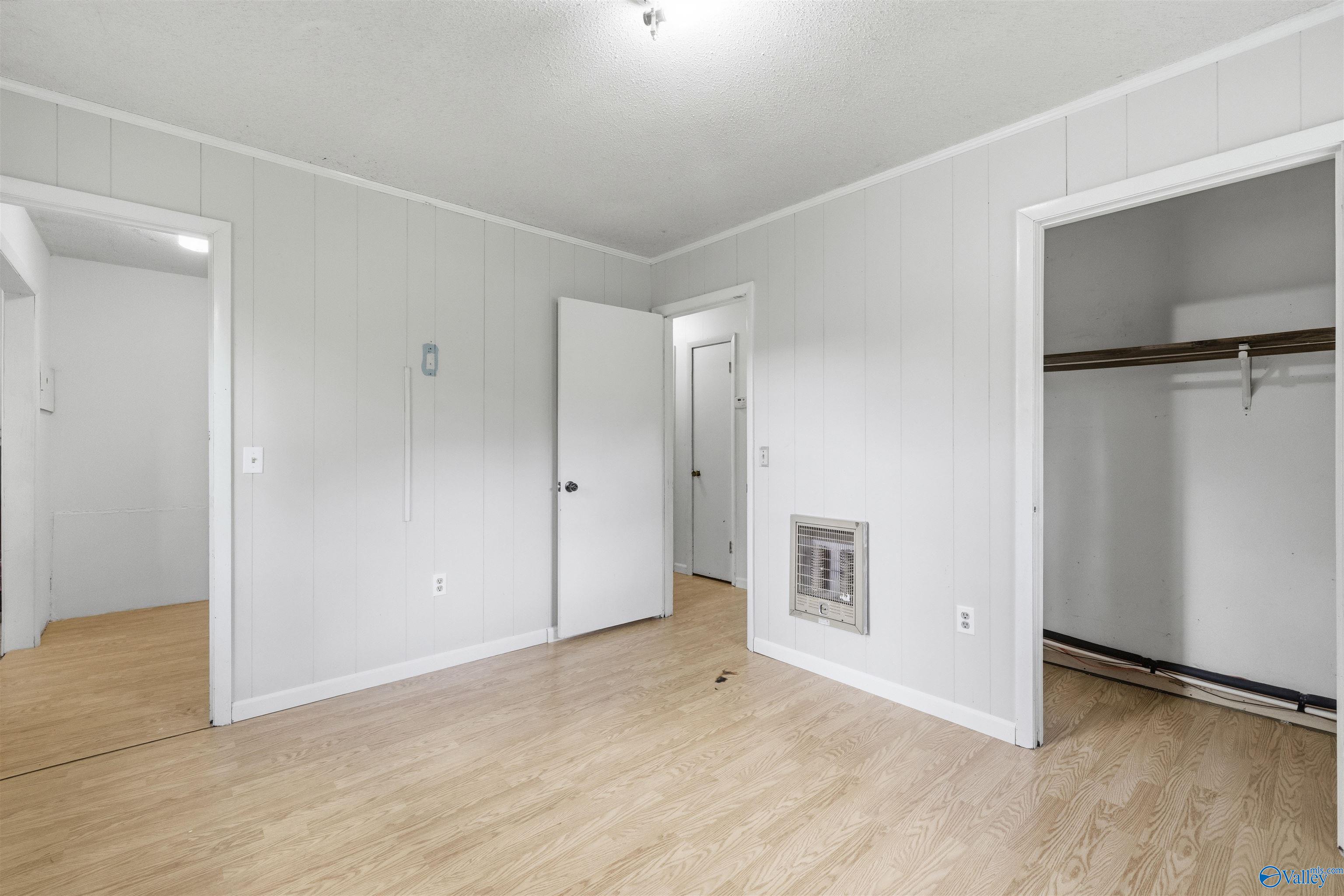Empty room with light wood flooring, white paneled walls, a wall heater, and an open closet.
