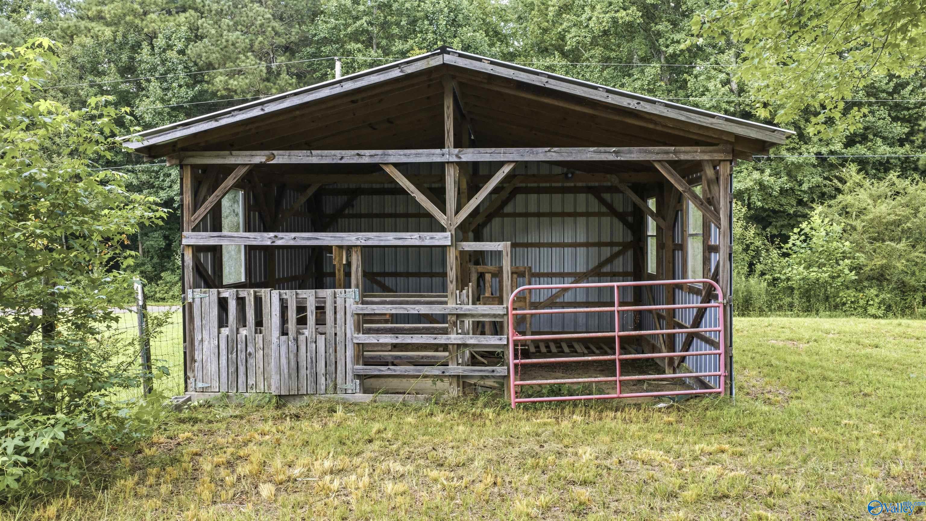 Open-front timber barn with corrugated siding, wooden fencing, and a red metal gate in a wooded clearing.