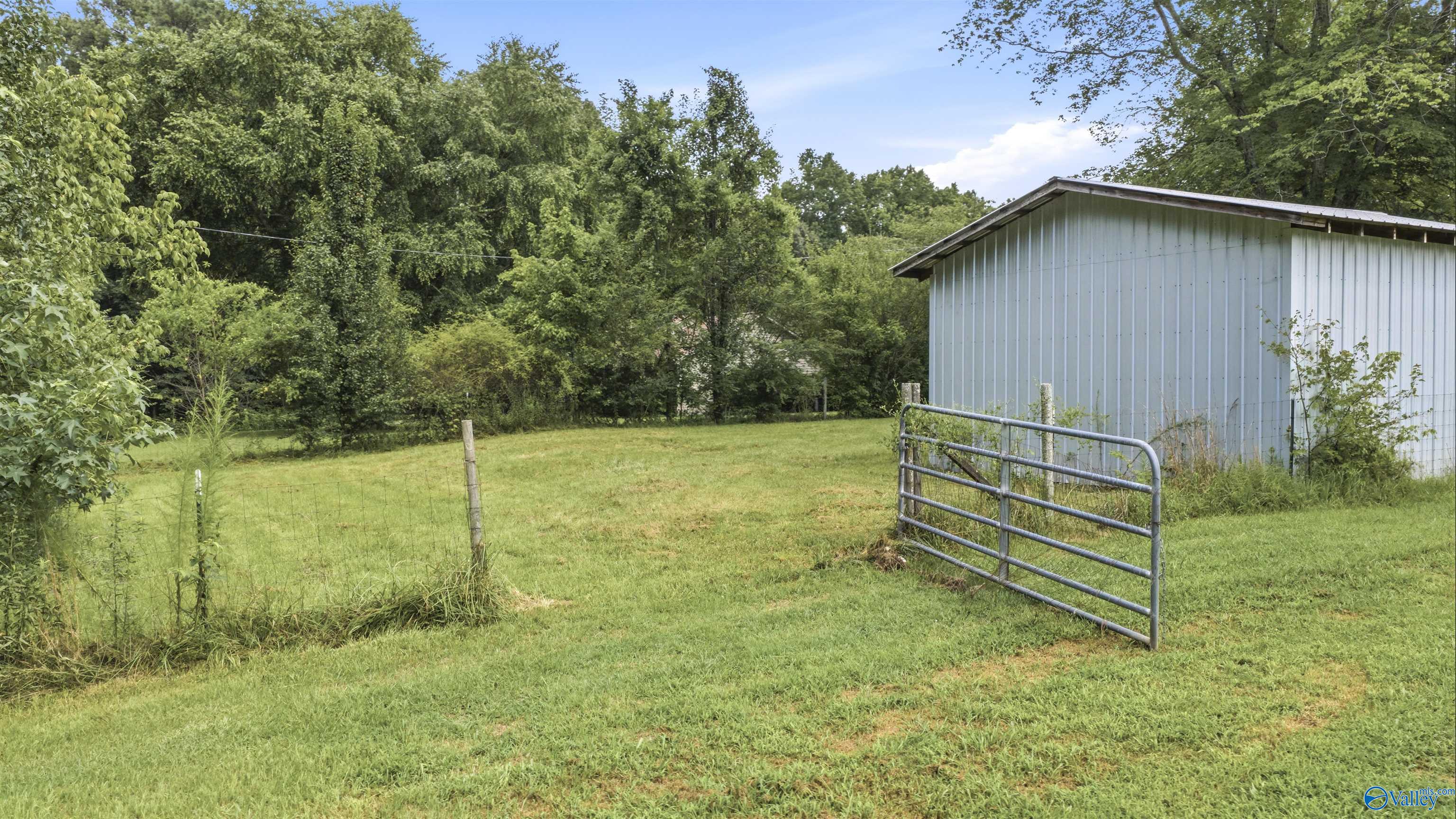 Fenced grassy yard beside a light blue metal outbuilding, with a metal gate and wooded surroundings.