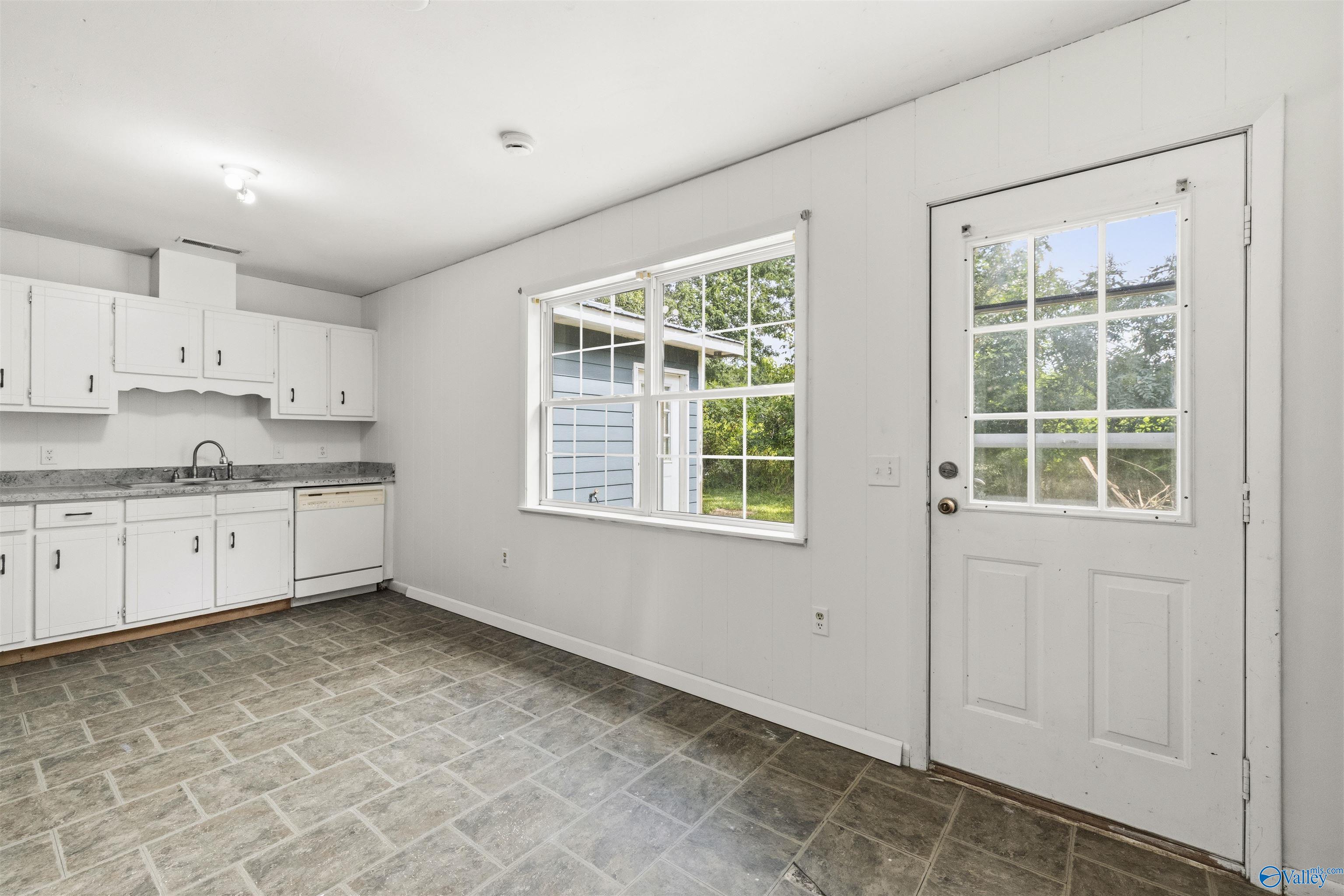 Vacant kitchen with white cabinets, gray tile flooring, and a window beside a glass-panel exterior door.