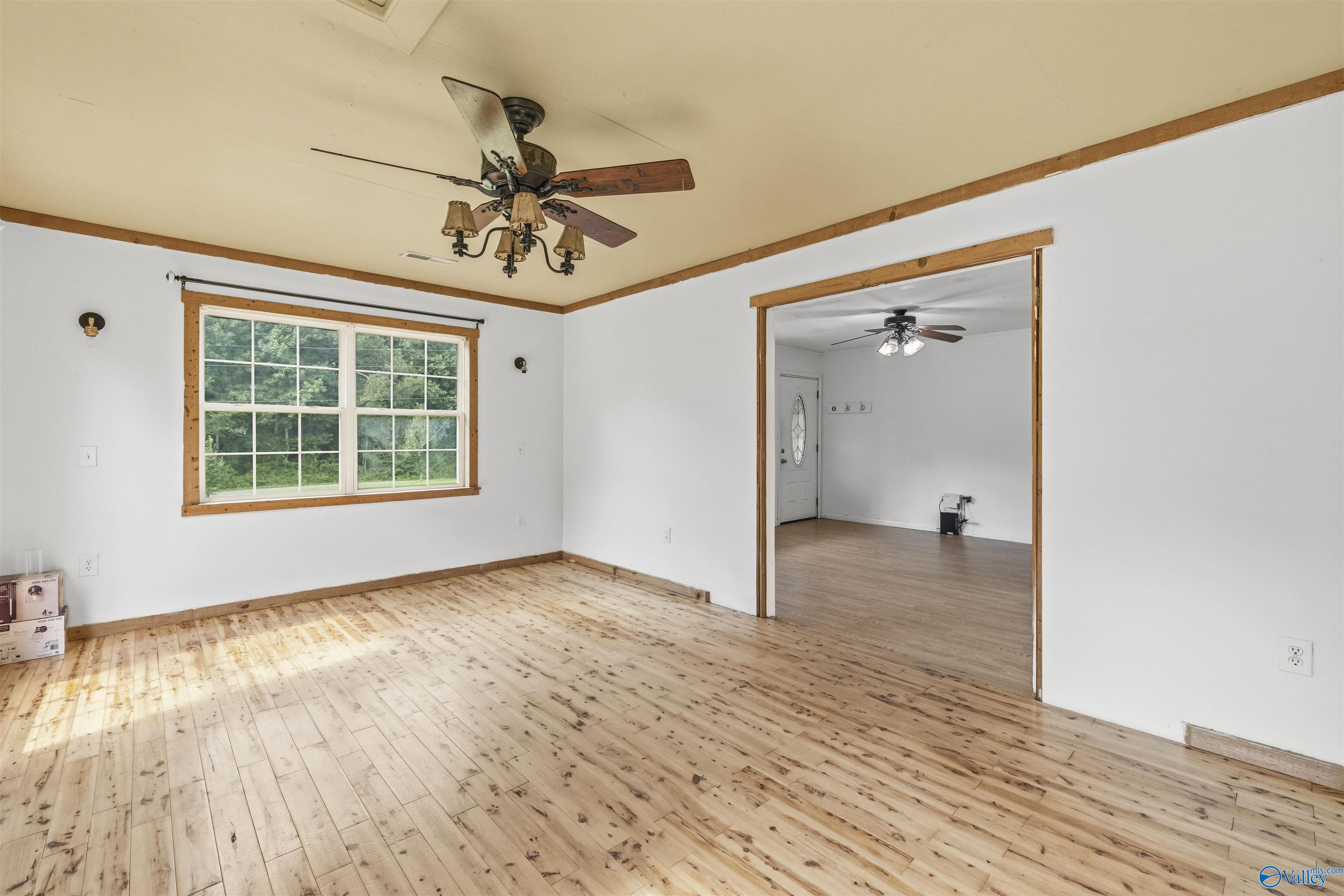 Vacant room with pale wood floors, a large window overlooking greenery, and an open doorway to an adjoining room.