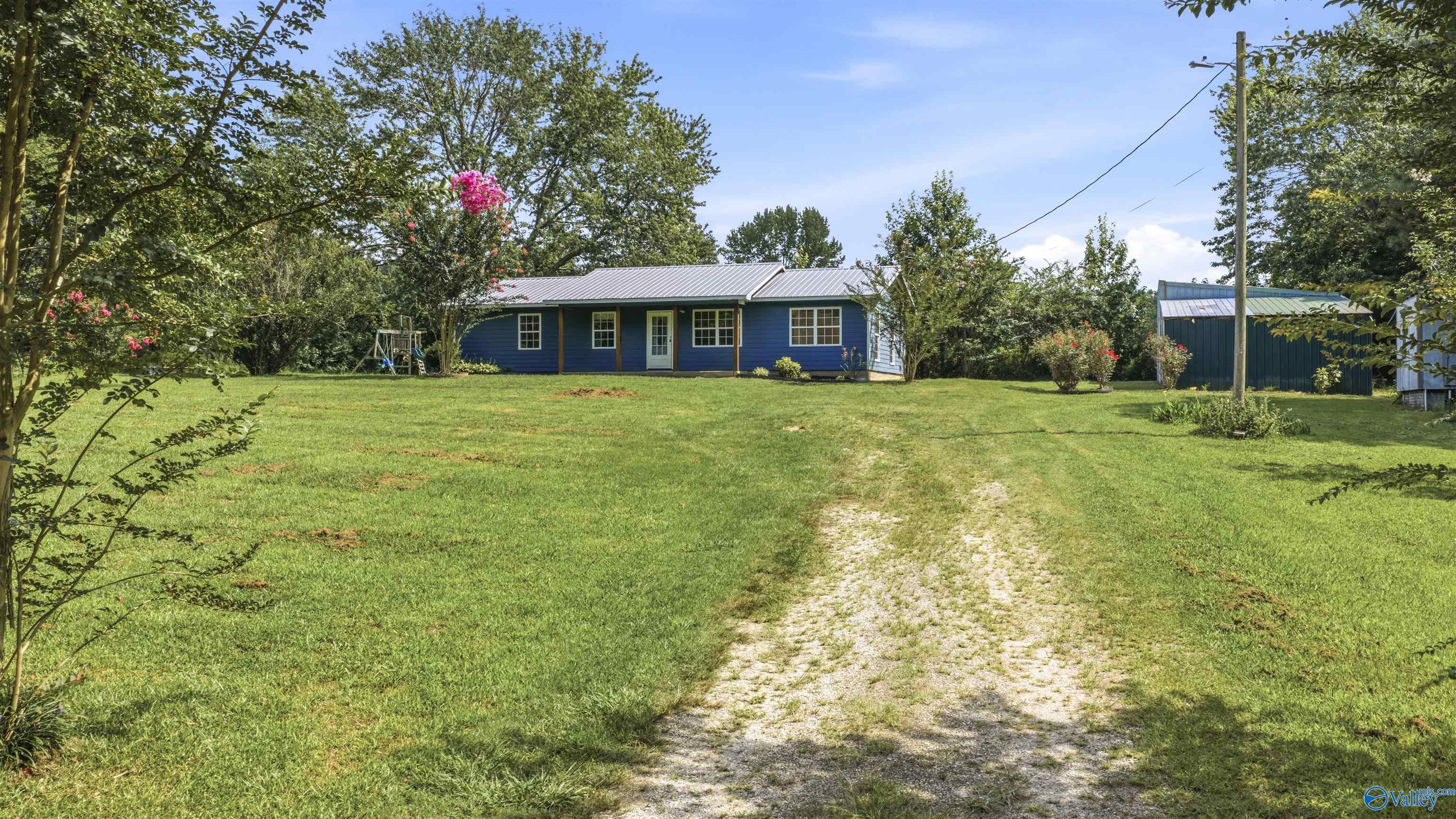 Blue single-story house beyond a broad lawn and gravel driveway, with a green metal outbuilding.
