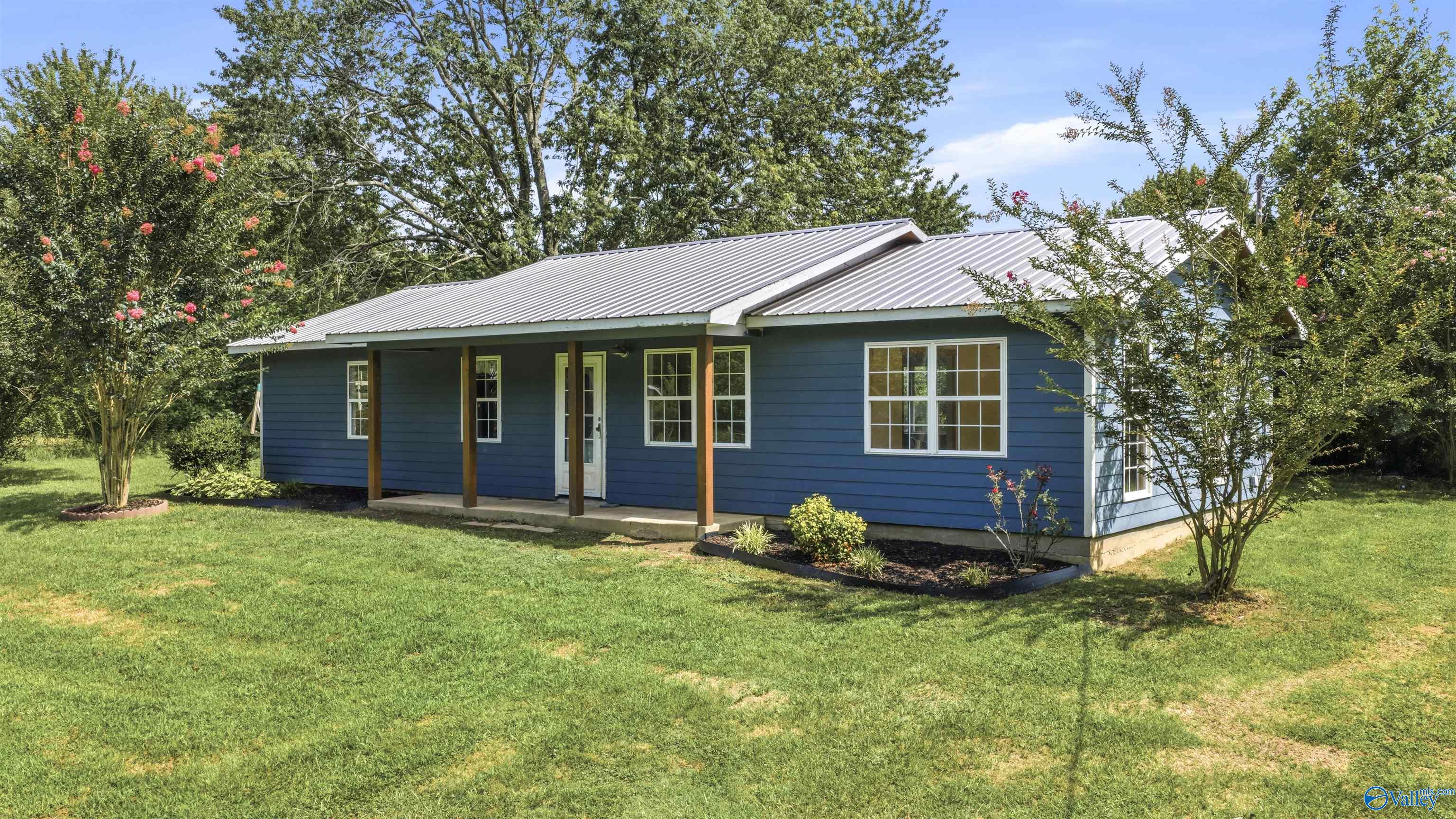 Blue home with a metal roof and covered porch, surrounded by a lawn and flowering shrubs.