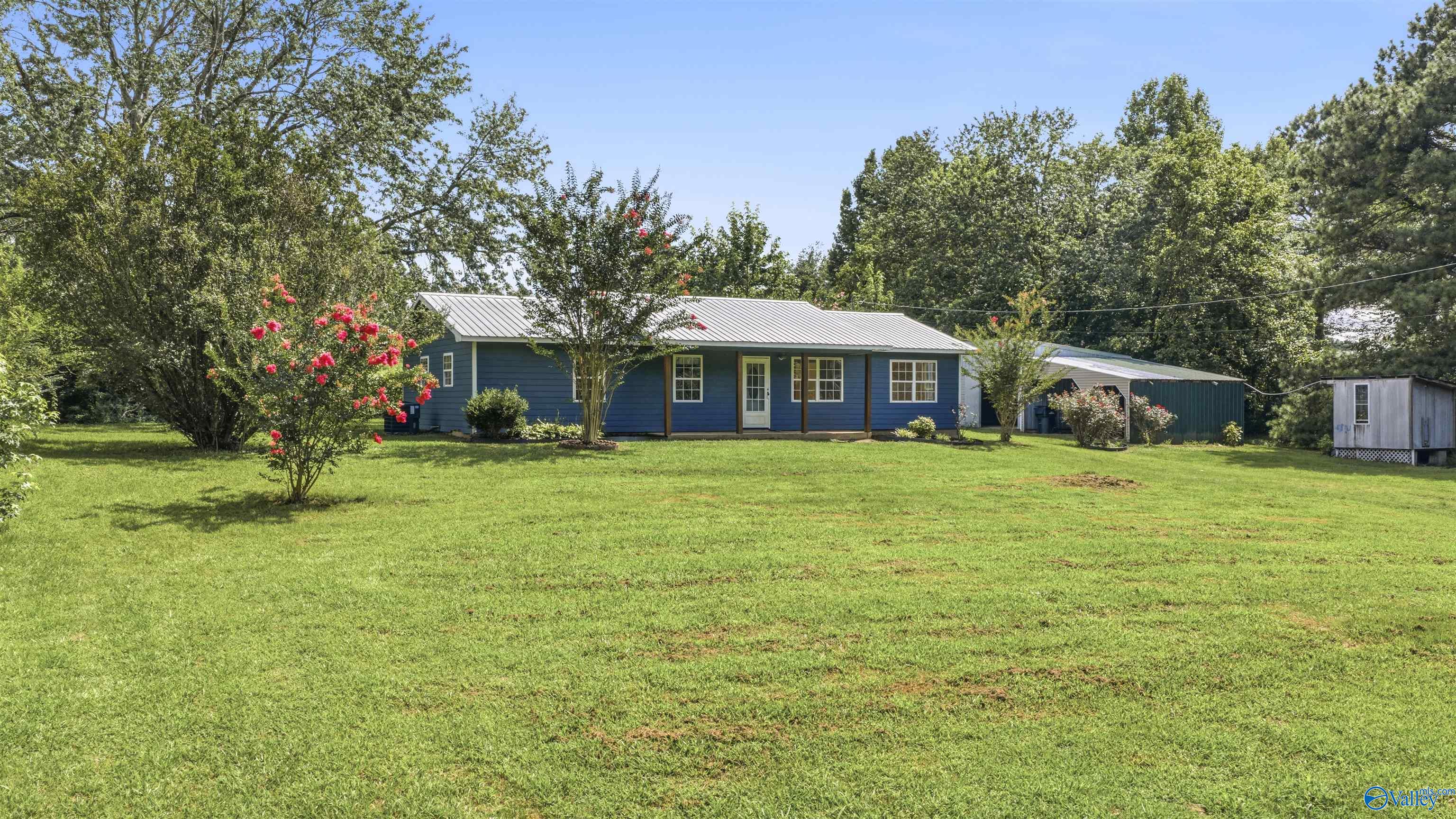 Blue single-story home with a covered porch, flowering shrubs, broad lawn, and two outbuildings.