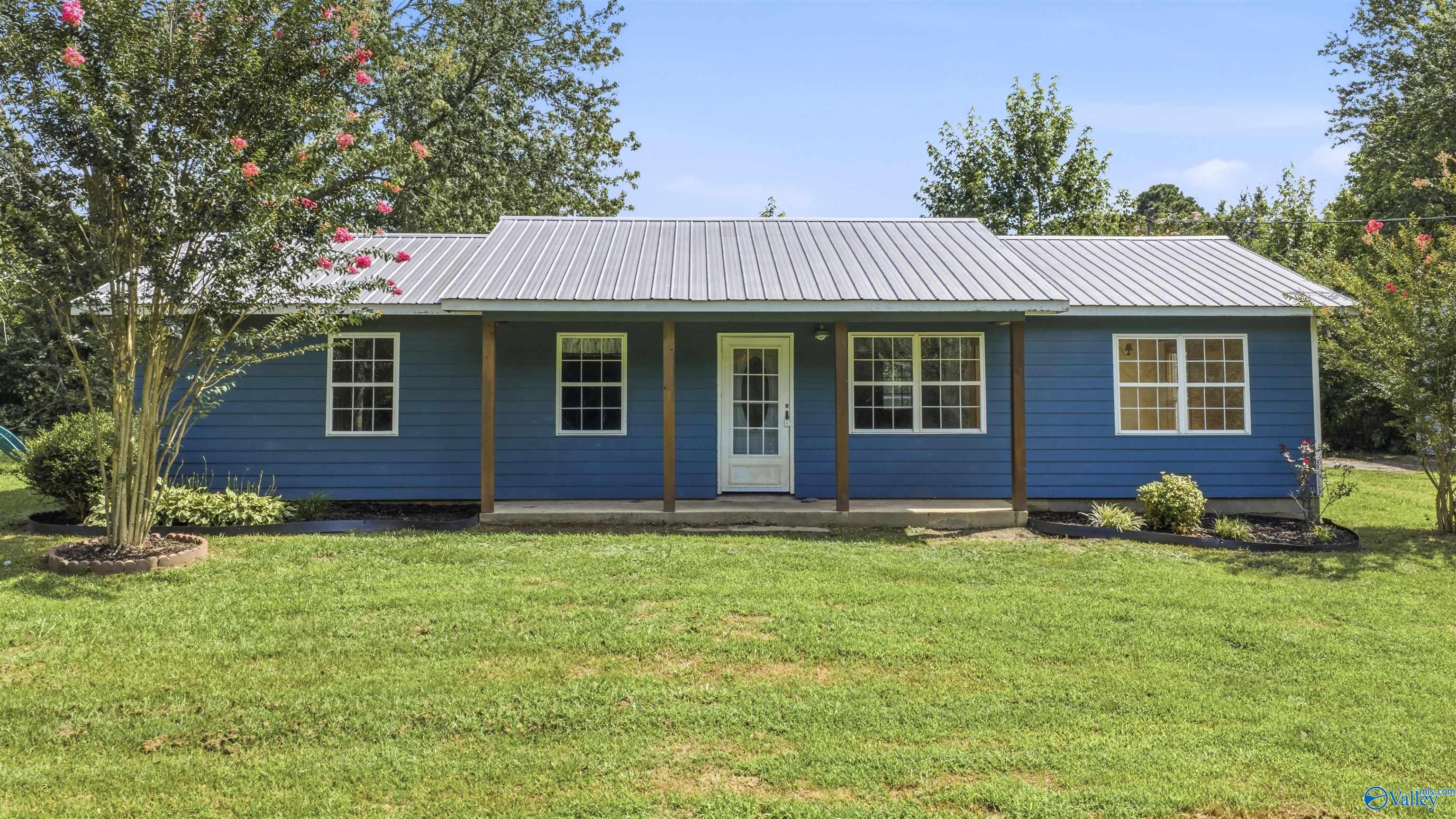 Blue single-story home with a covered porch, metal roof, and flowering trees beside the lawn.