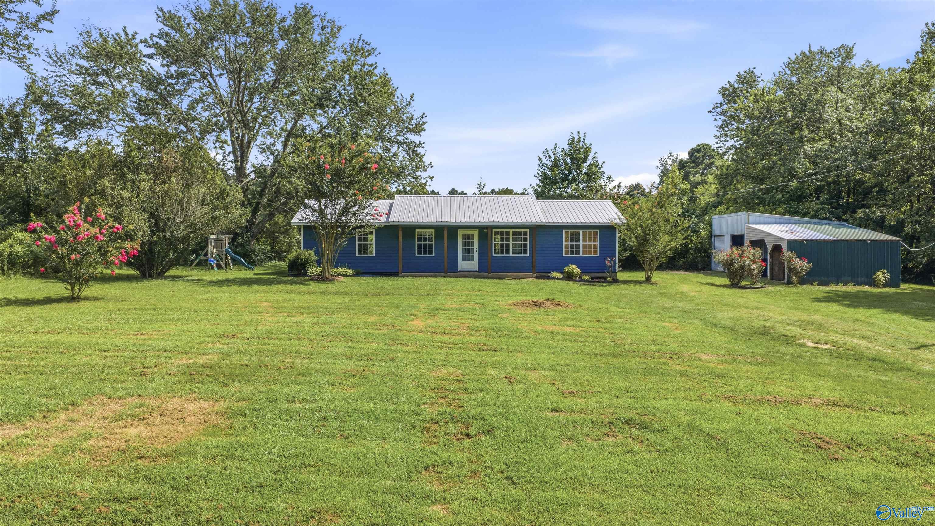 Blue single-story home with a metal roof, broad lawn, flowering shrubs, playset, and detached outbuilding.