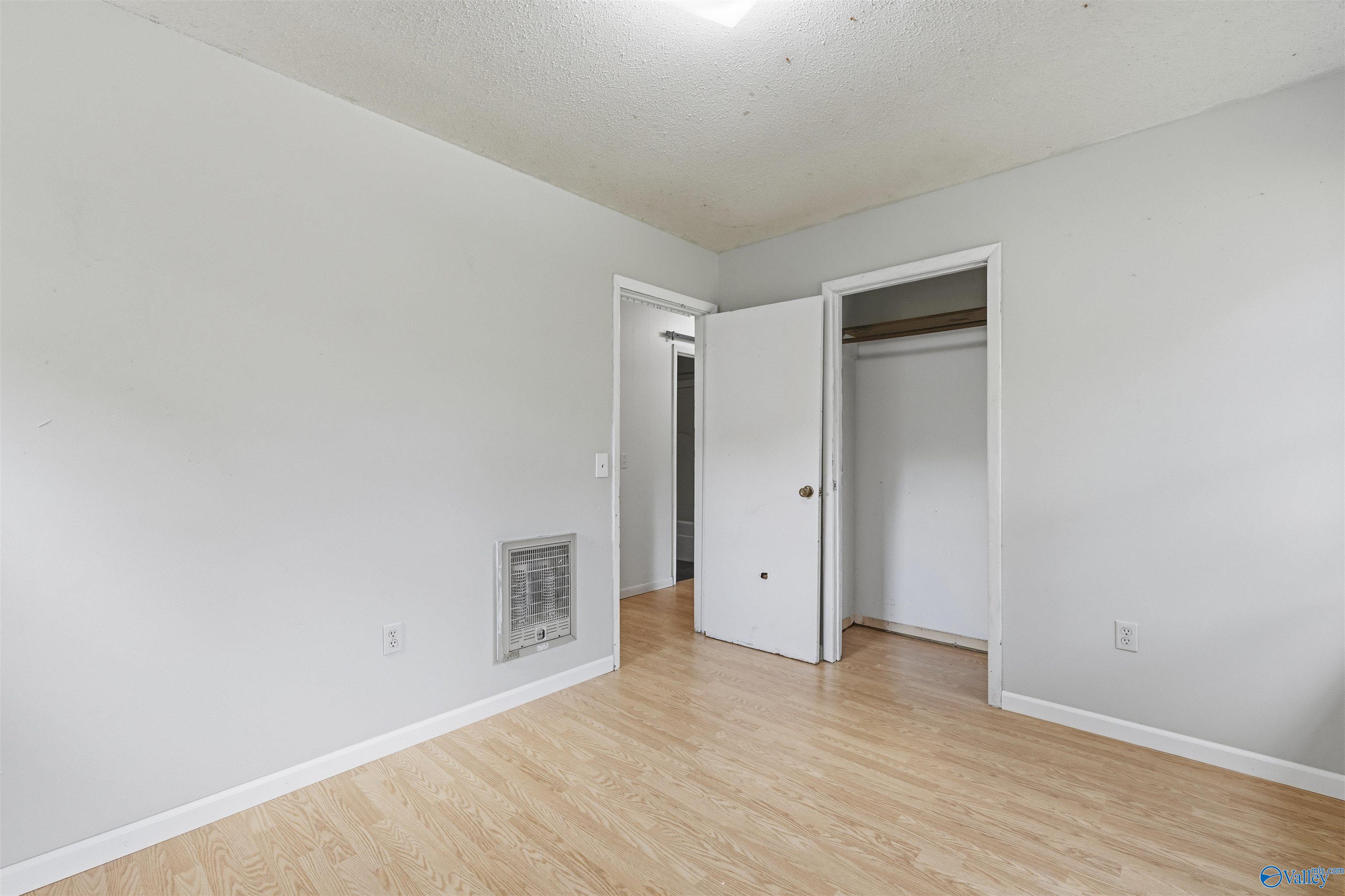 Empty bedroom with light wood flooring, wall heater, and open closet.