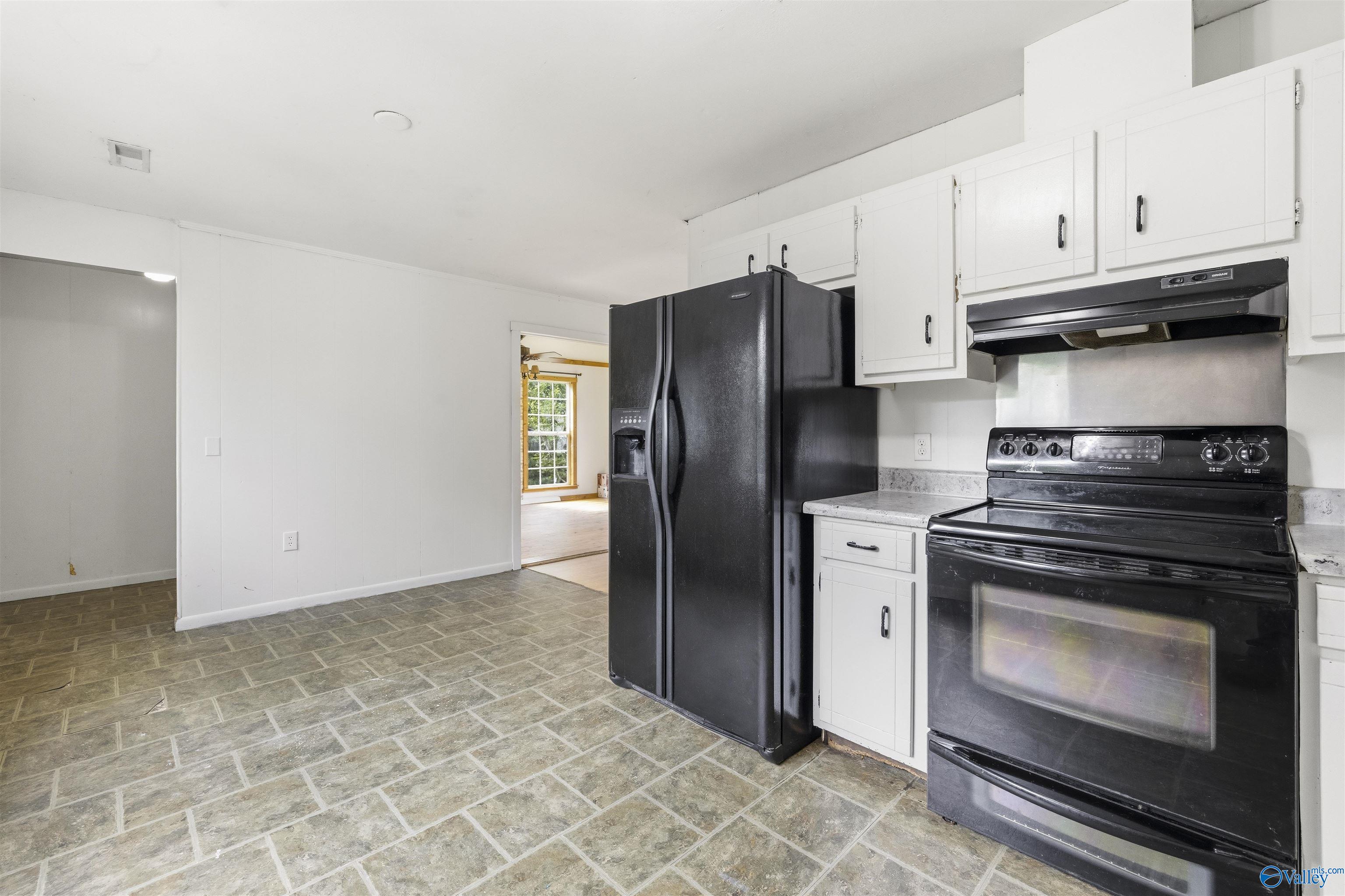 Kitchen with white cabinets, black appliances, gray tile flooring, and a doorway to a room with a window.