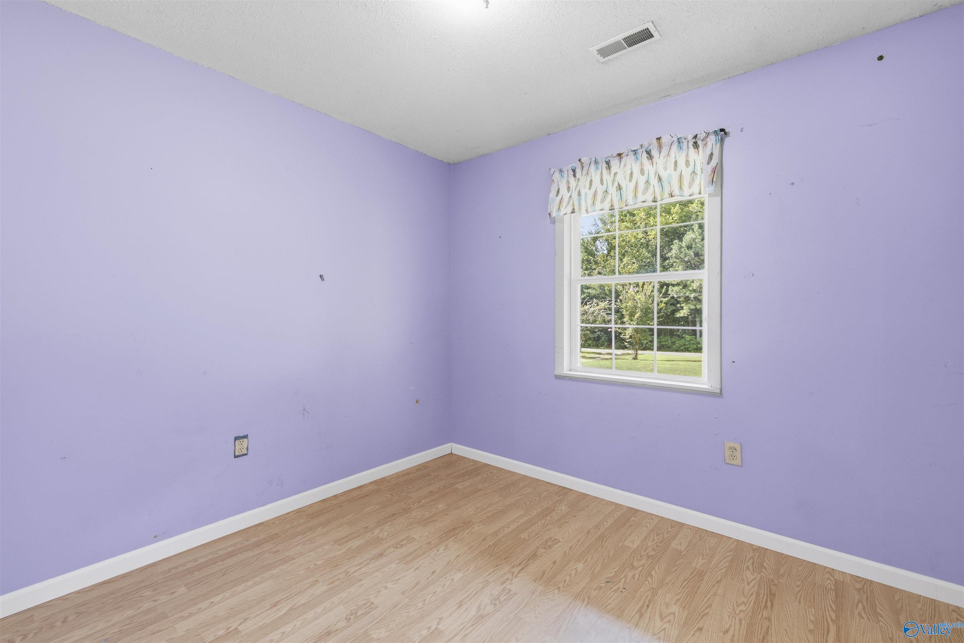 Empty bedroom with lavender walls, wood-look flooring, and a window overlooking a green yard.