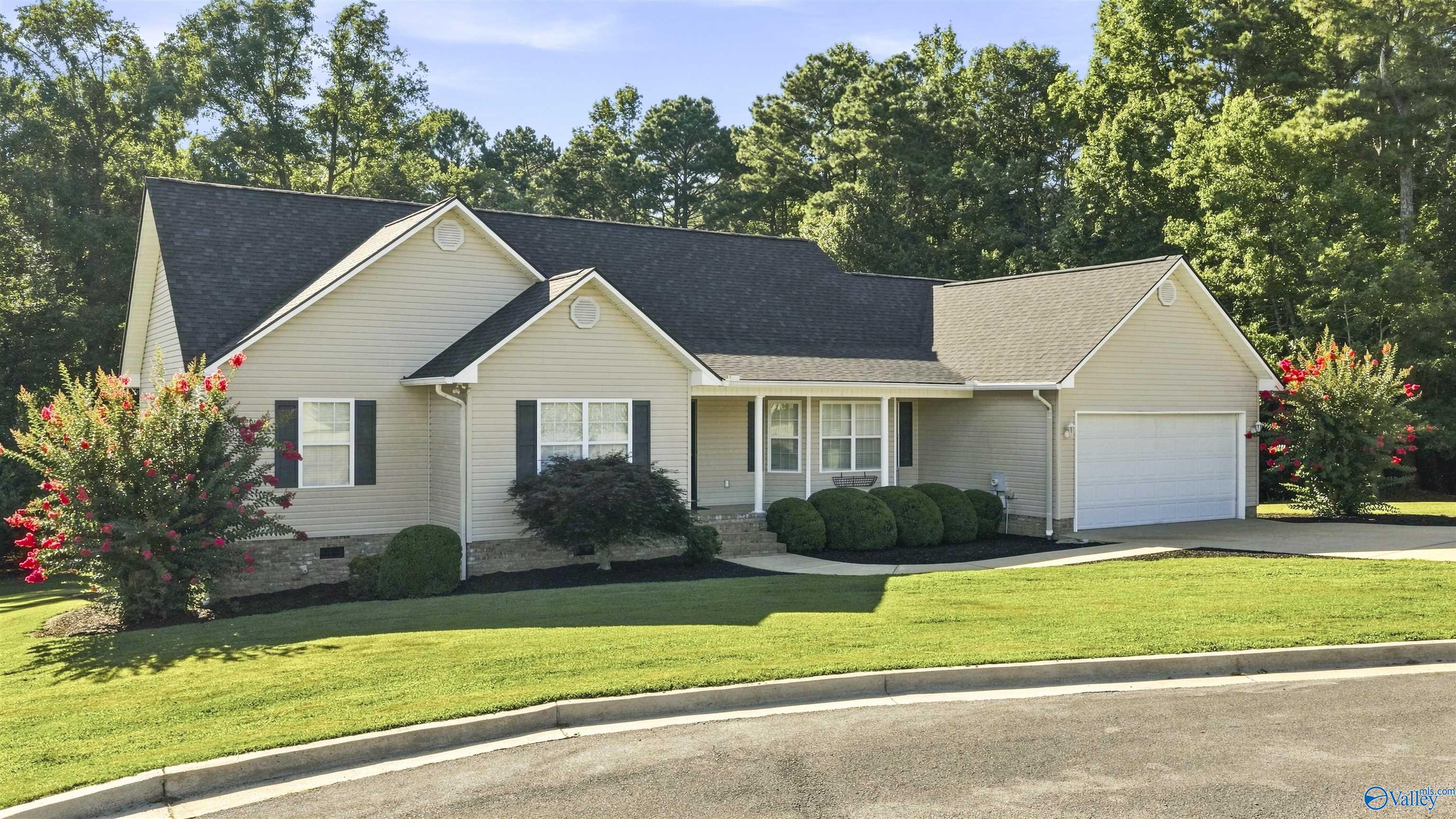 Beige gabled home with an attached garage, manicured lawn, and flowering shrubs.