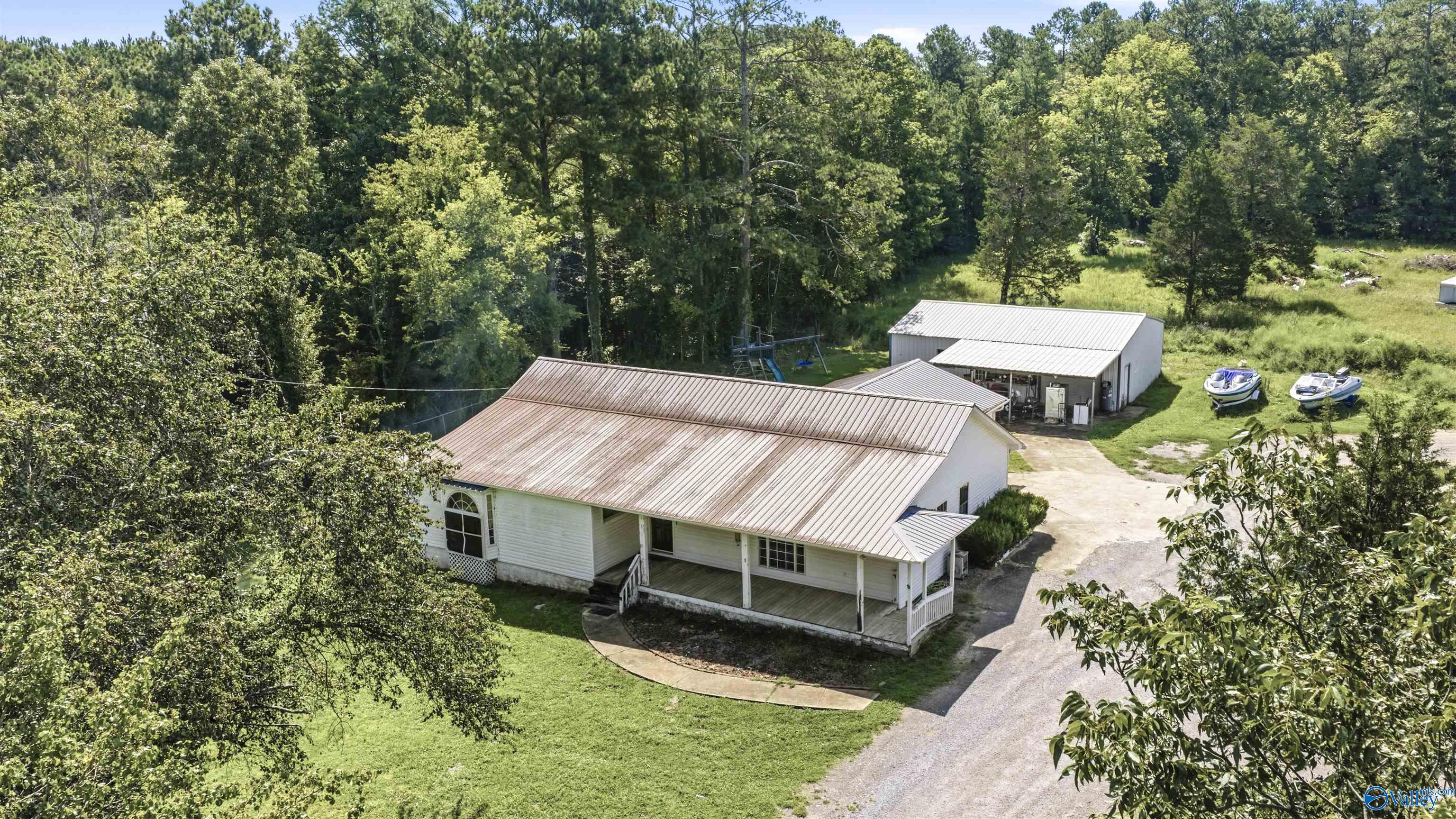 Aerial view of a white home with a covered porch, outbuilding, and two boats among trees.