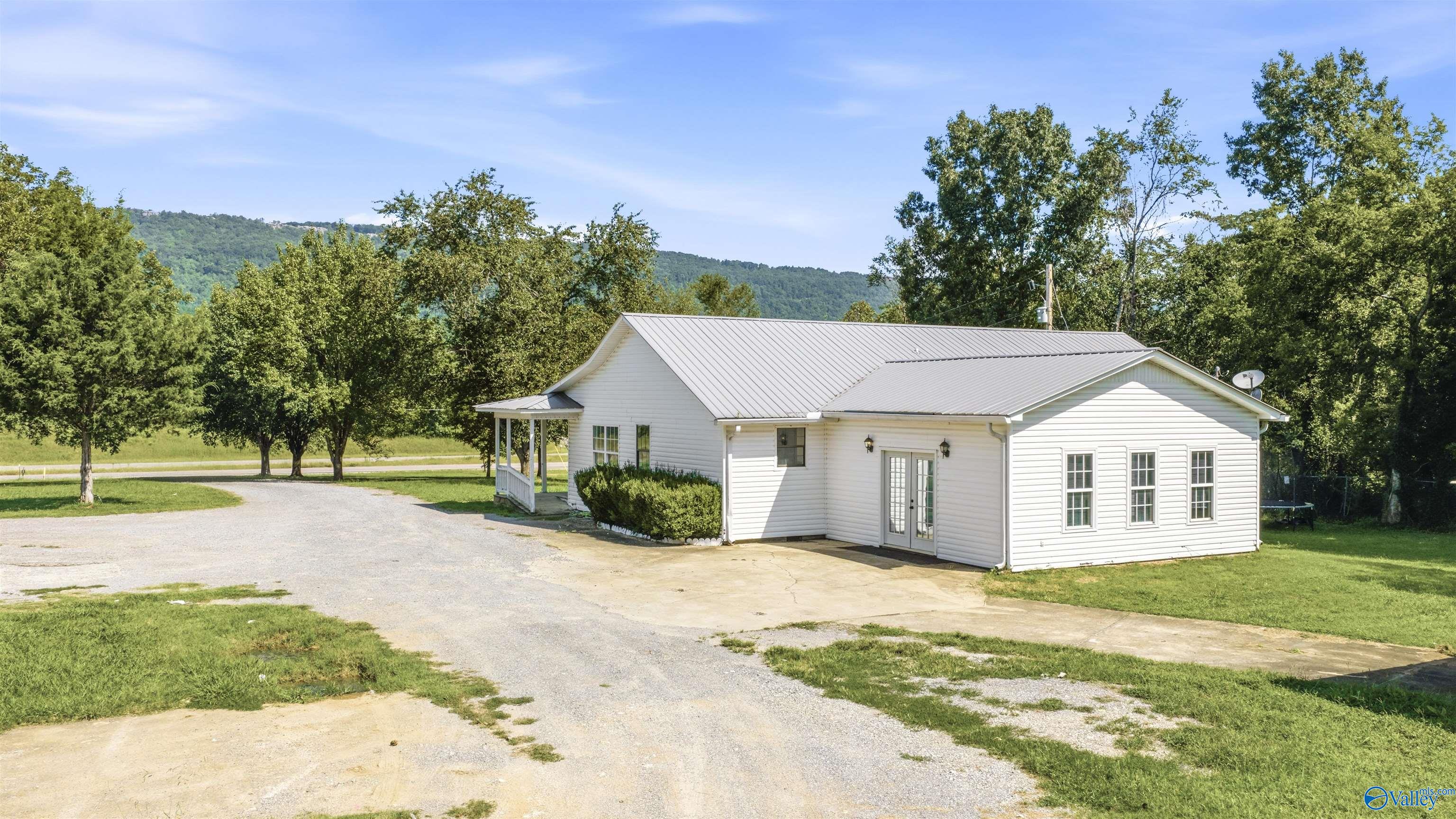 White-sided home with a light metal roof, gravel driveway, and wooded hills behind it.