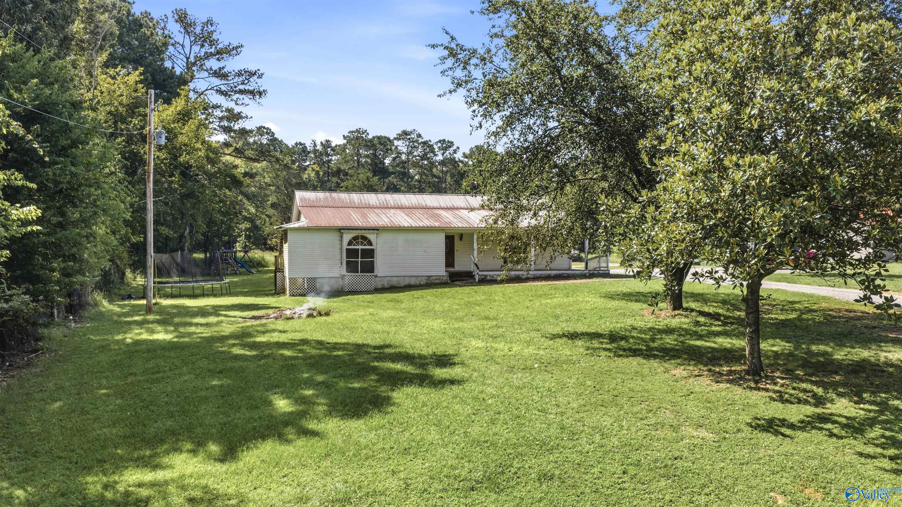 Single-story white home with a metal roof behind a spacious lawn and mature trees, with a trampoline beside it.