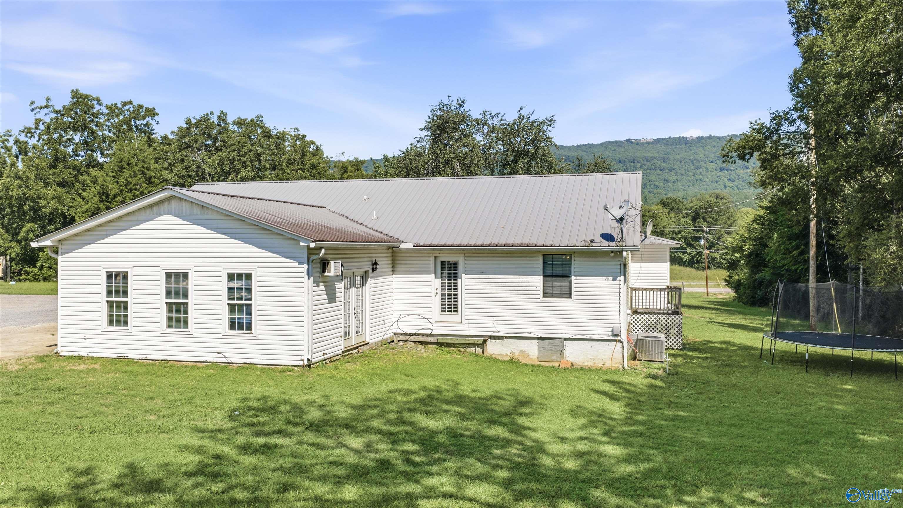 White-sided home with a metal roof, broad lawn, trampoline, and wooded mountain backdrop.