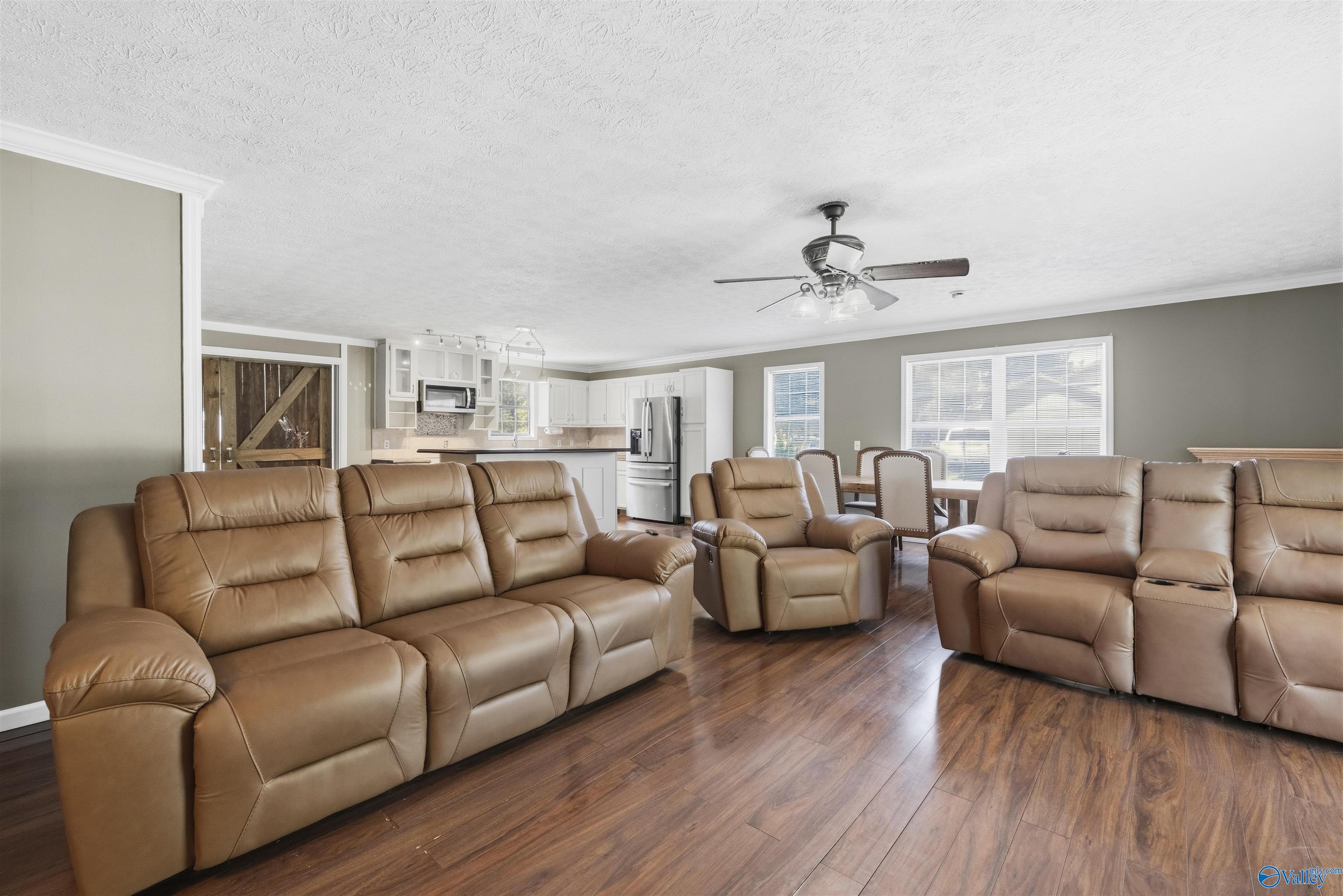Open-plan living room with tan leather recliners, dark wood-look floors, and a kitchen and dining area beyond.
