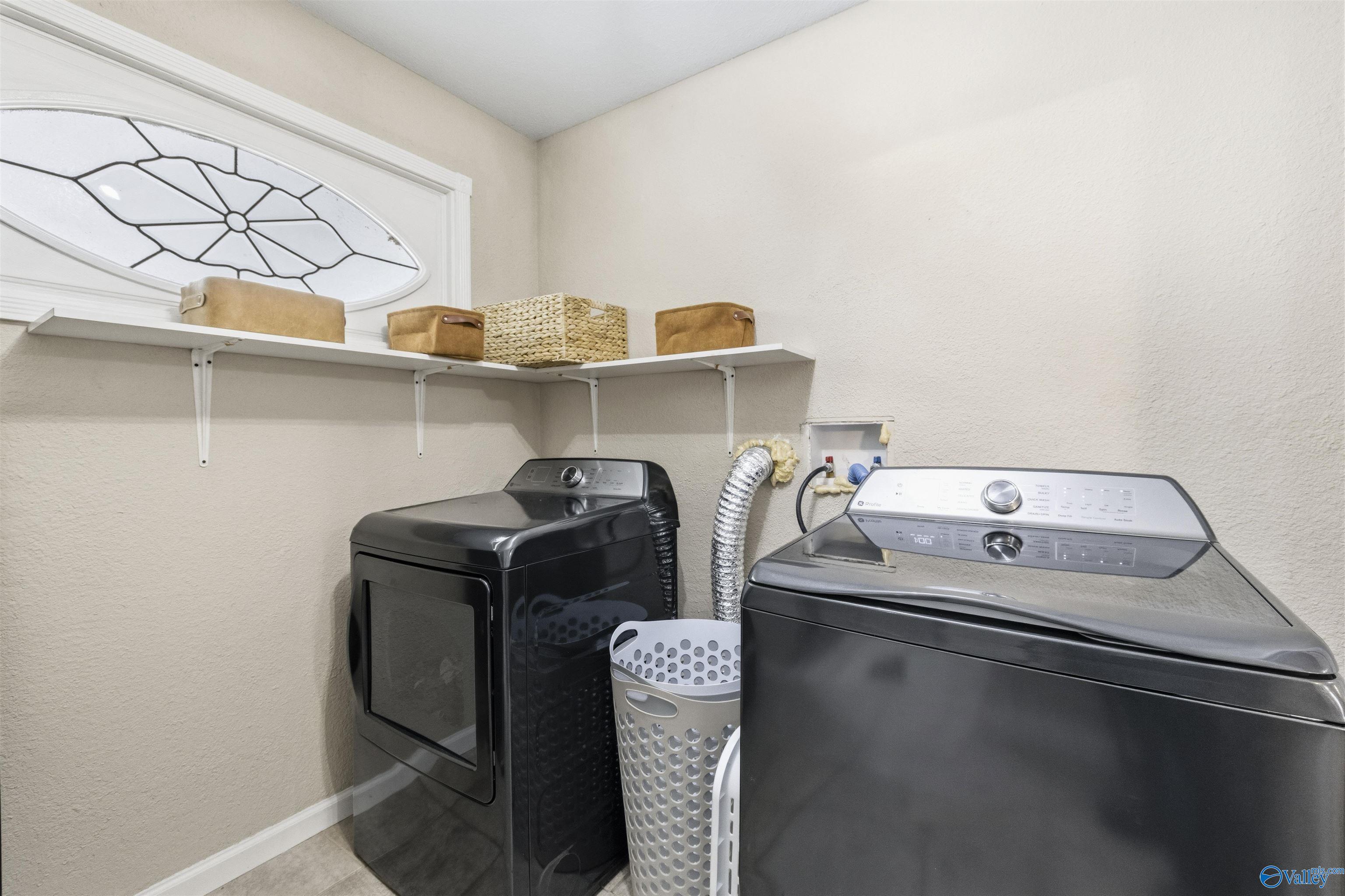 Laundry room with dark washer and dryer beneath wall shelves, beside an oval decorative glass panel.