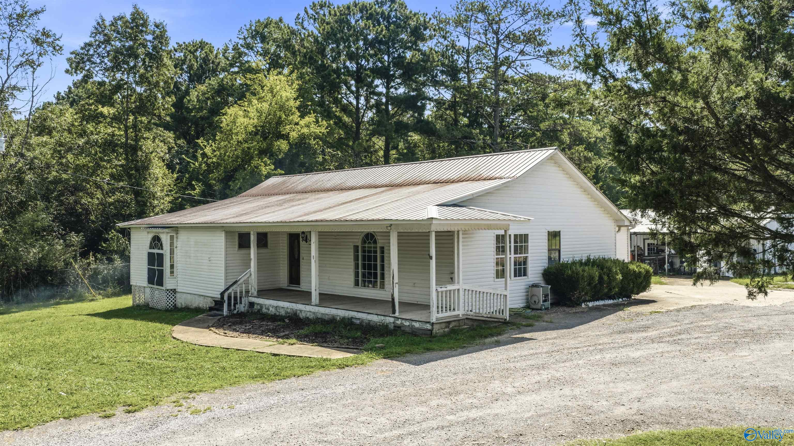 White single-story home with a metal roof, covered porch, and gravel driveway amid mature trees.