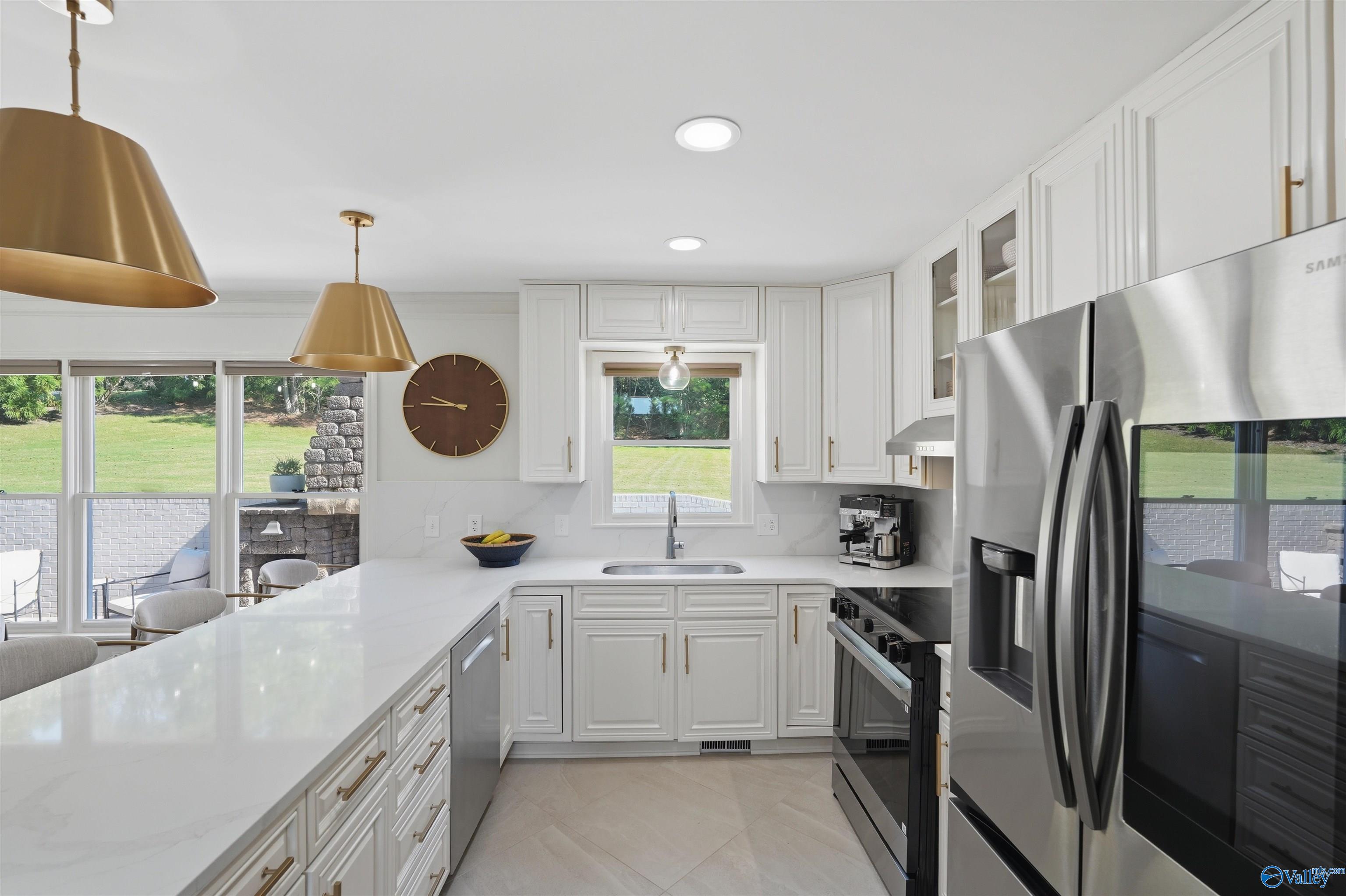 Bright white kitchen with stainless steel appliances, brass pendant lights, and large windows overlooking the yard.